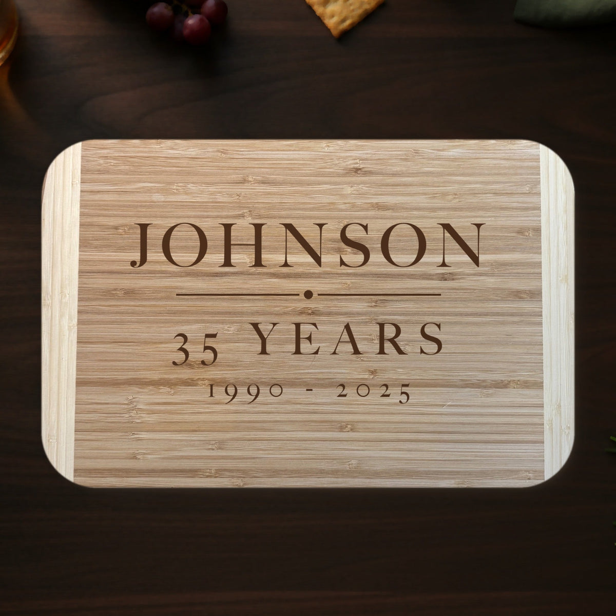Personalized retirement cutting board on a dark walnut kitchen counter with grapes, crackers and whiskey, engraved JOHNSON 35 YEARS 1990 to 2025 on two tone bamboo.