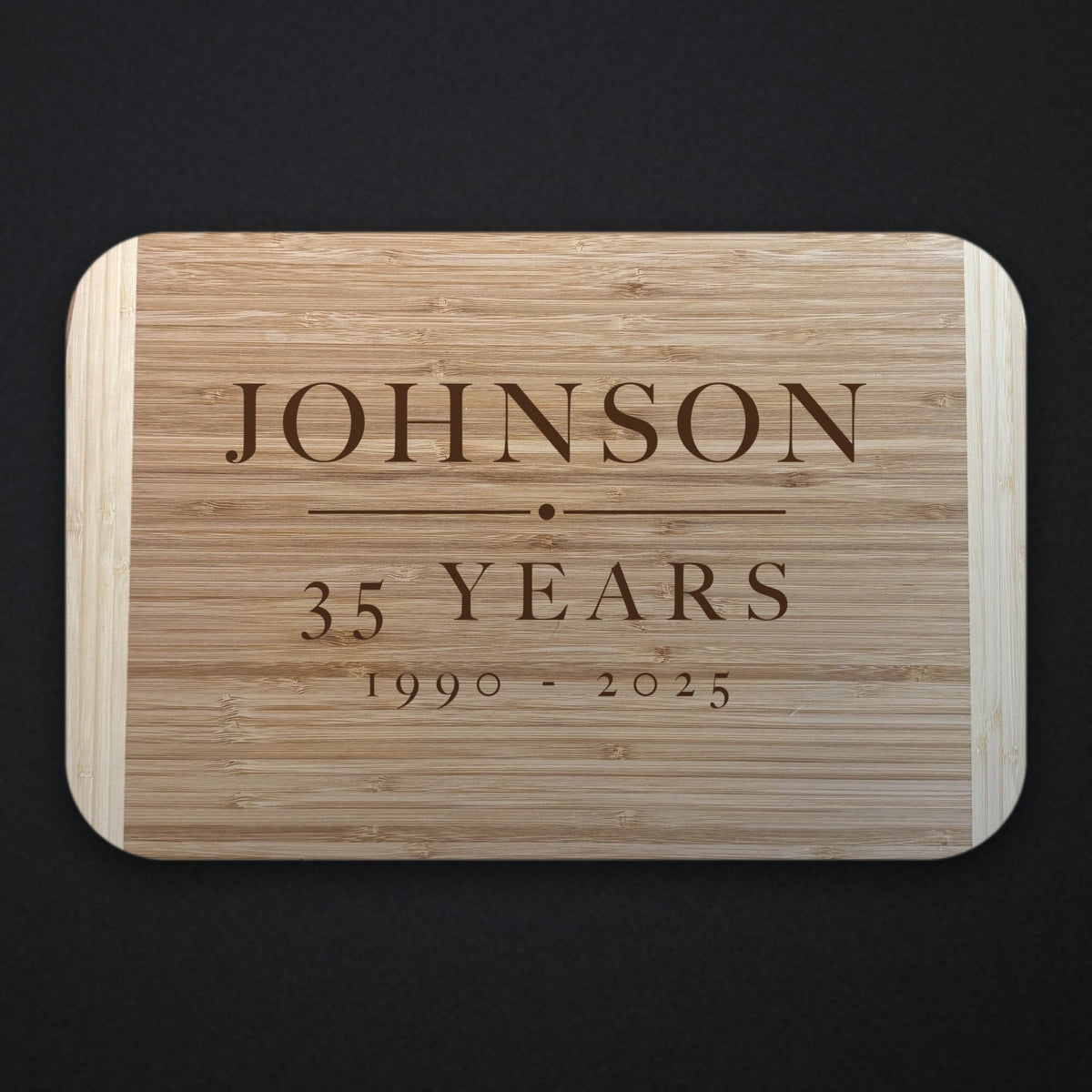 Two tone bamboo retirement cutting board engraved with the name JOHNSON above a rule, with 35 YEARS and the dates 1990 to 2025 beneath, shown on a dark background.
