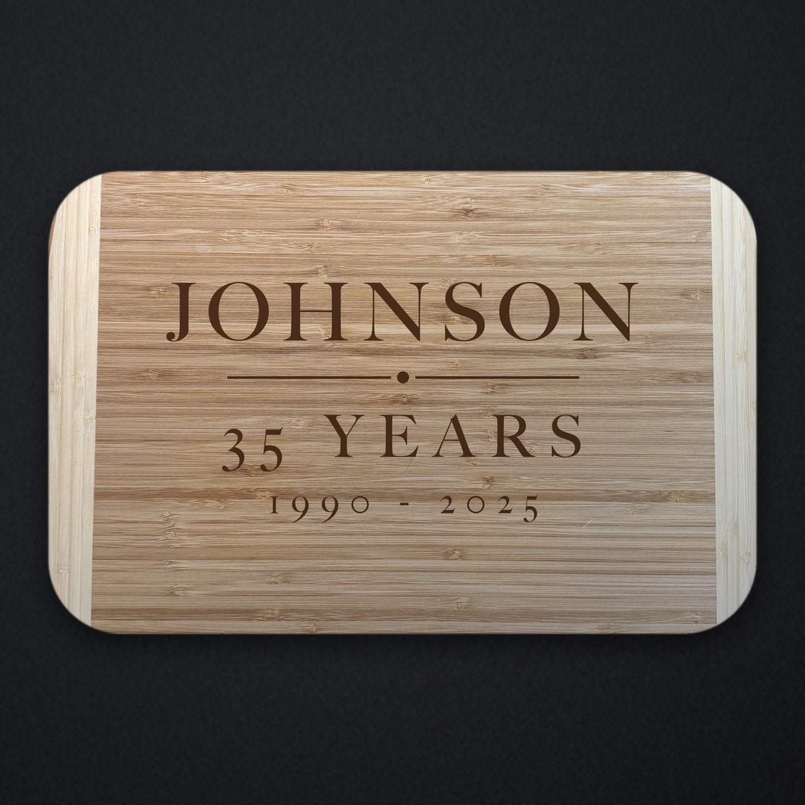 Two tone bamboo retirement cutting board engraved with the name JOHNSON above a rule, with 35 YEARS and the dates 1990 to 2025 beneath, shown on a dark background.