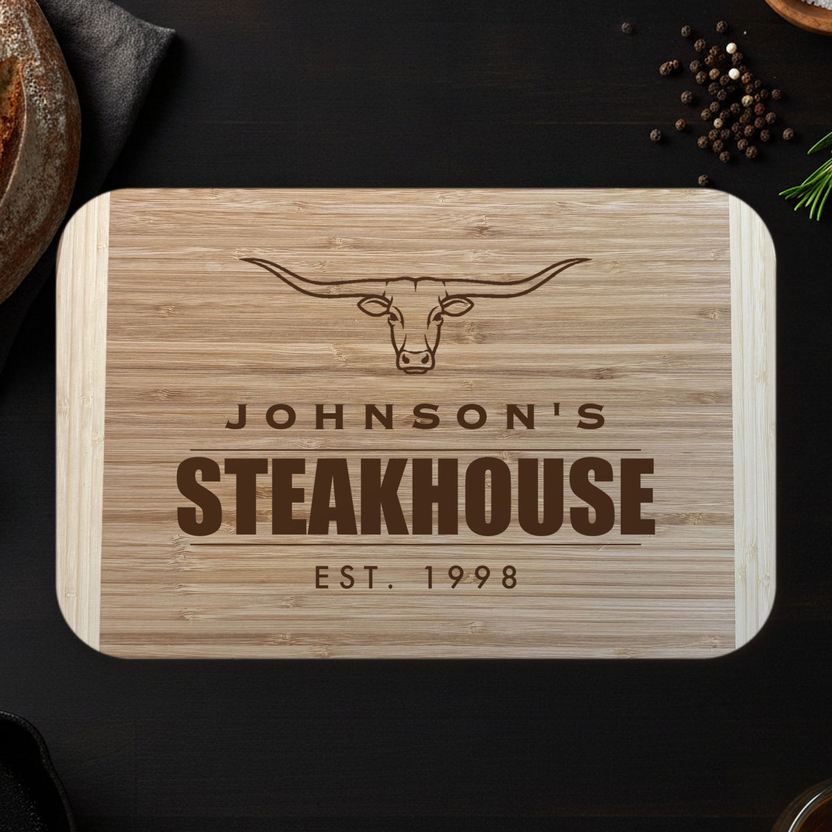 Personalized steakhouse cutting board on a dark espresso counter with bread and rosemary, engraved JOHNSON'S STEAKHOUSE EST. 1998 under a longhorn steer on two tone bamboo.