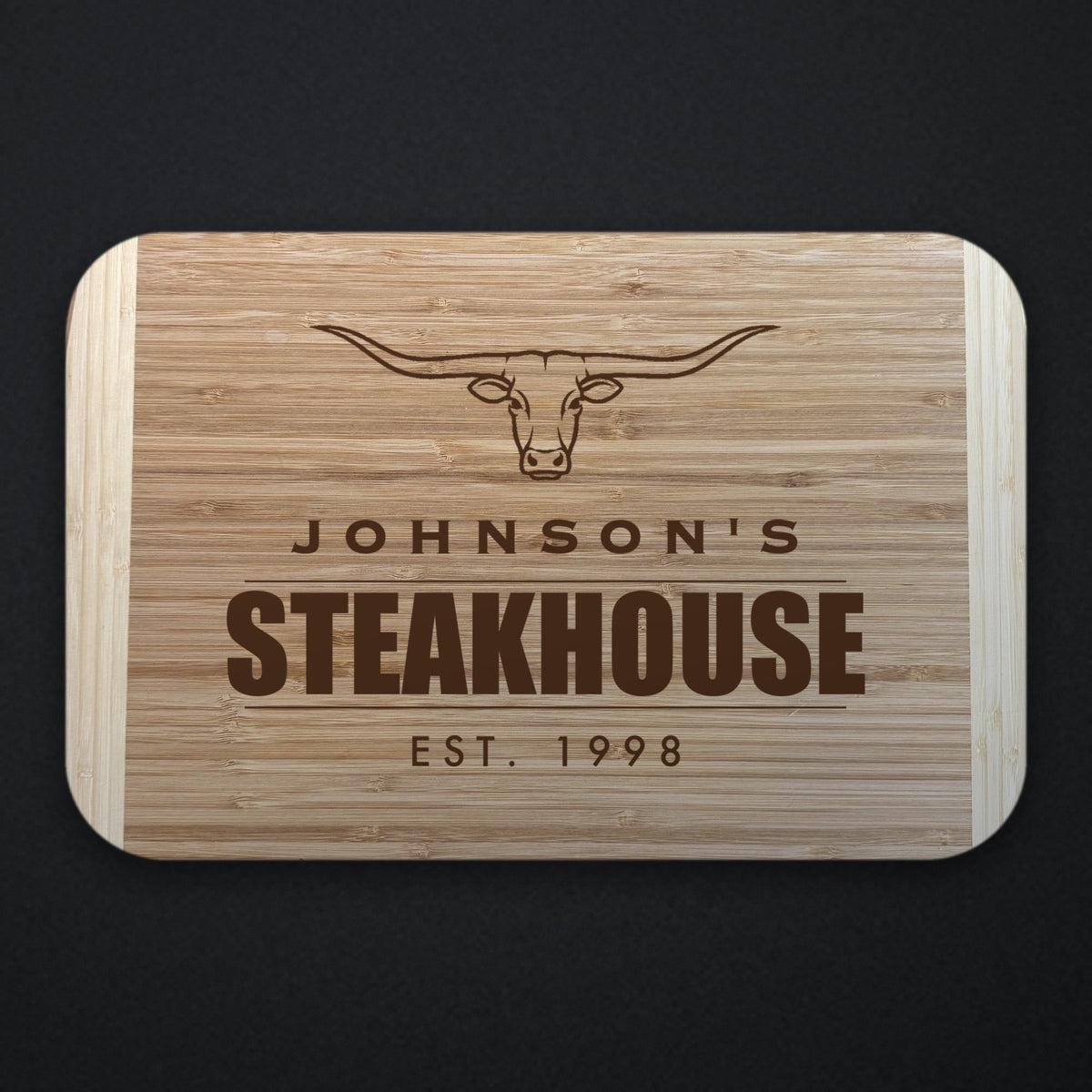 Two tone bamboo cutting board engraved with a longhorn steer above the name JOHNSON'S and the word STEAKHOUSE, with EST. 1998 below, shown on a dark slate background.