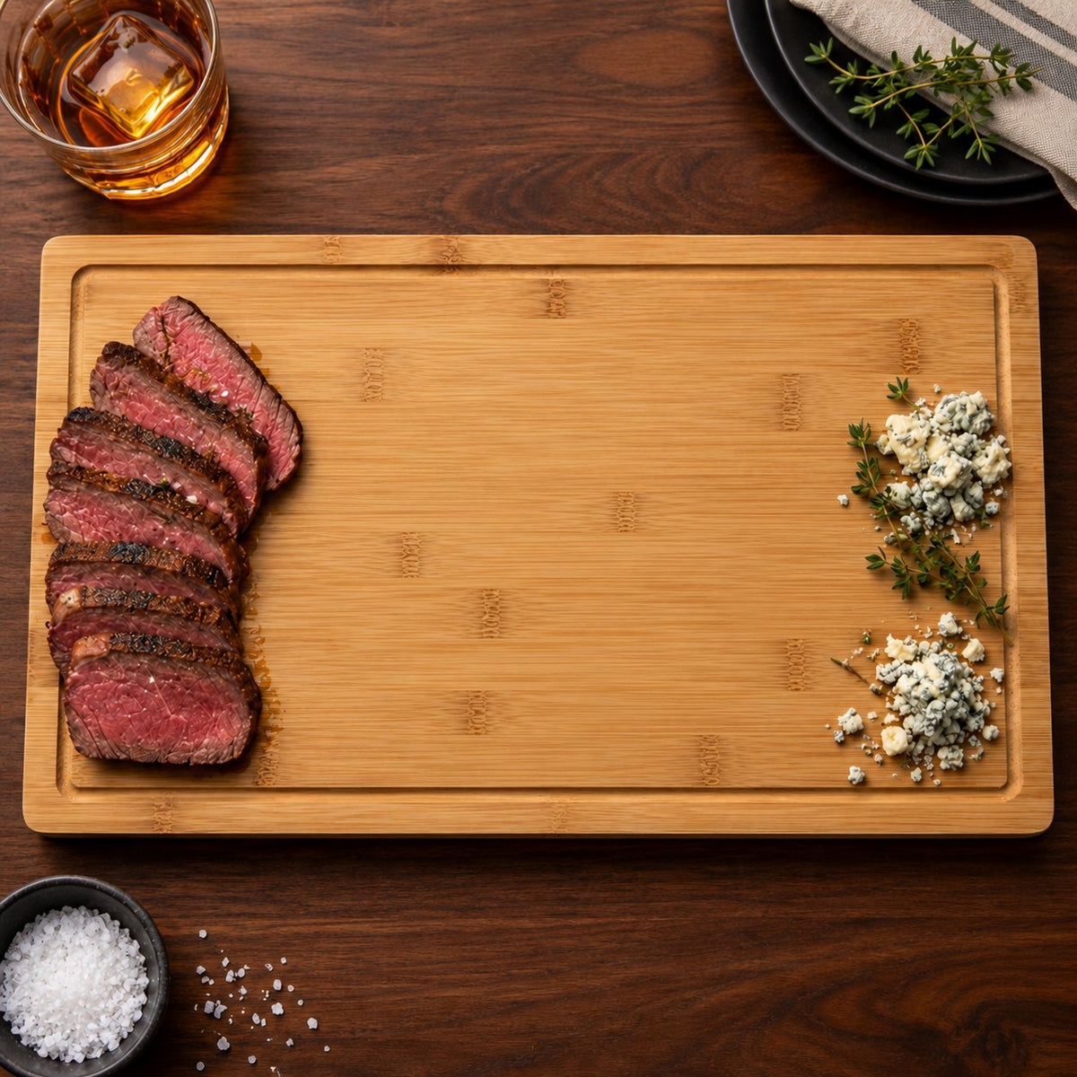 Bamboo steakhouse cutting board ready for his name, with sliced steak, blue cheese, a whiskey glass and dark plates on a walnut table