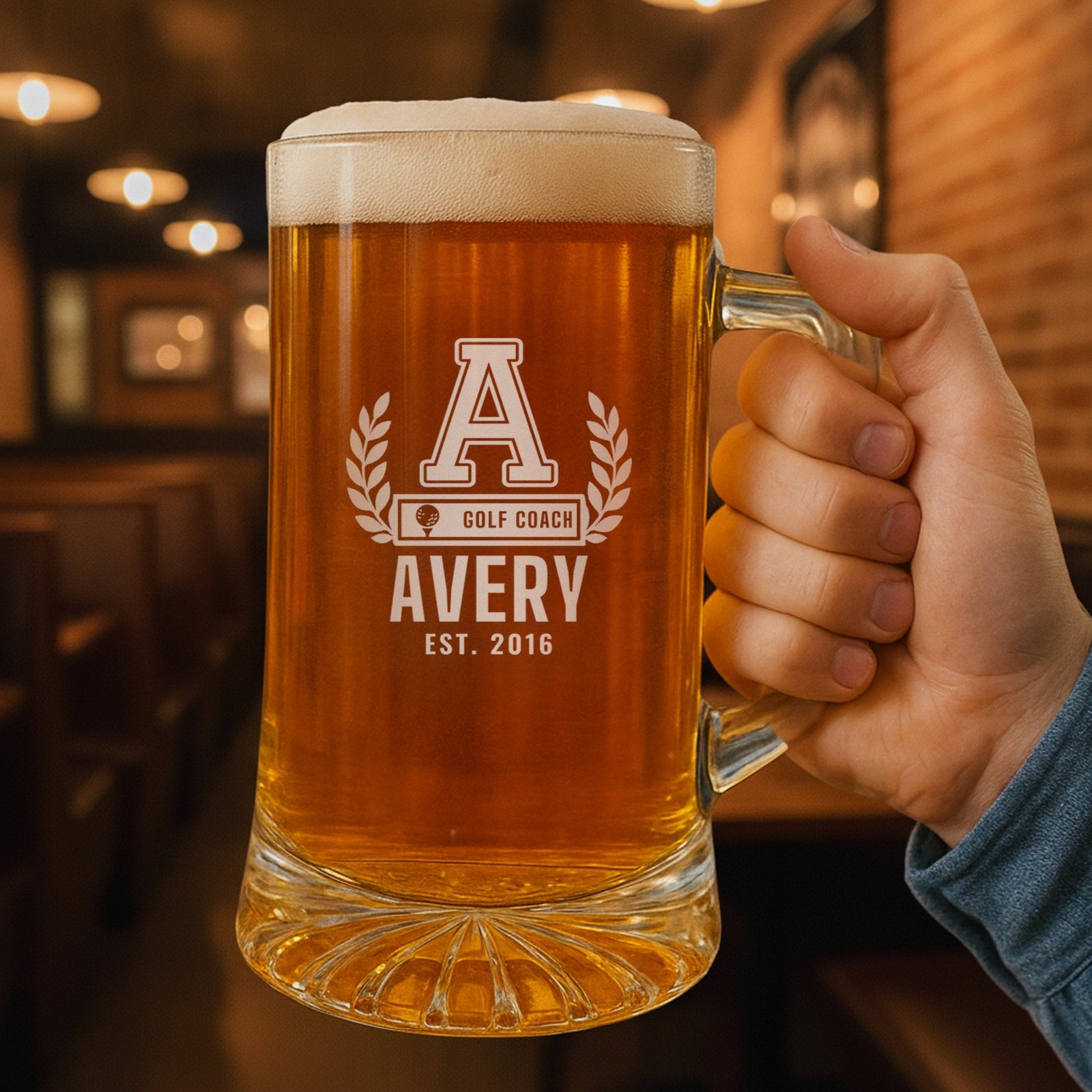 Hand holding the 25 oz personalized golf coach letterman beer stein in a pub, engraved with a varsity letter with laurel and a golf ball COACH bar: A, Golf Coach, Avery, Est. 2016
