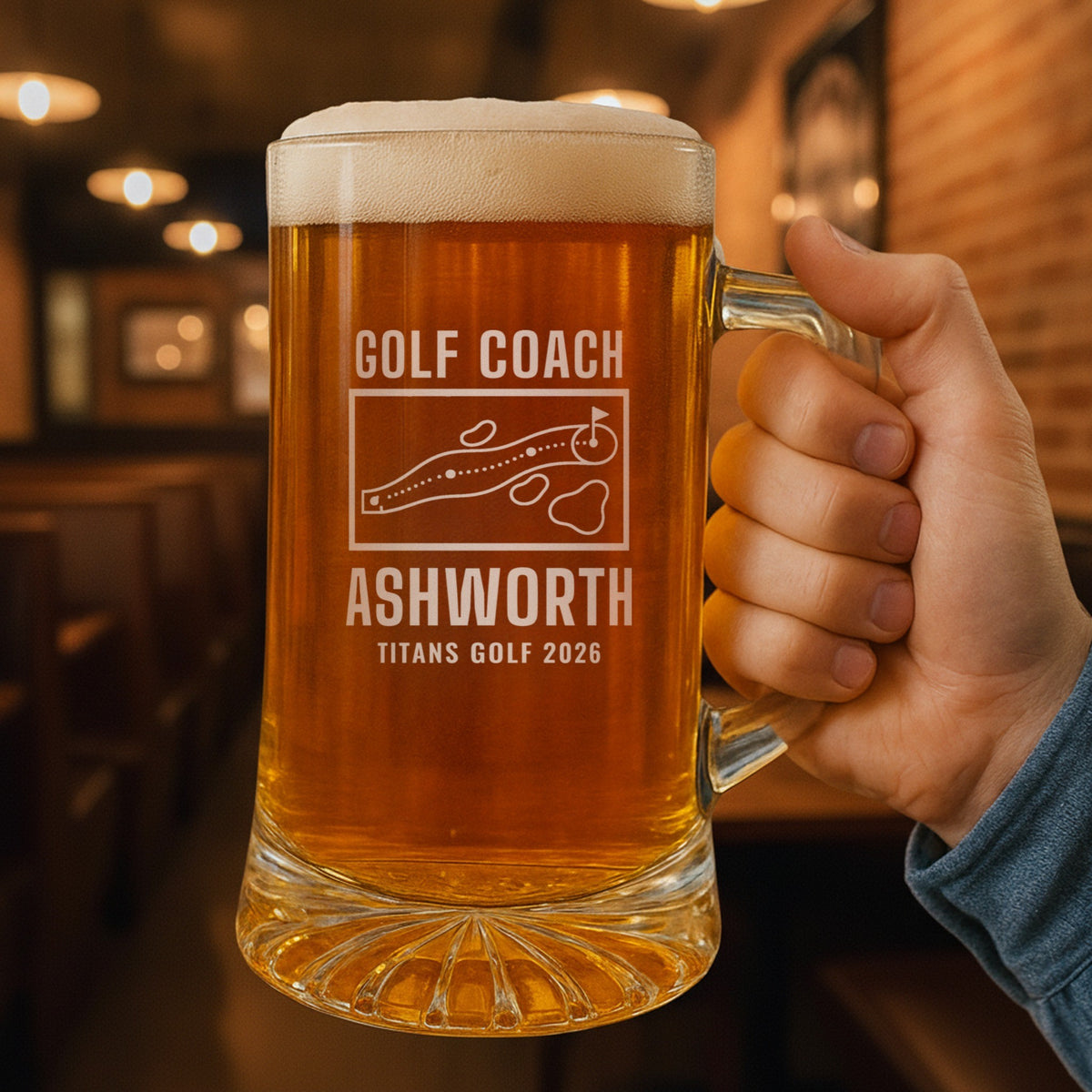 Hand holding the 25 oz personalized golf coach yardage book beer stein in a pub, engraved with a golf hole map with tee, fairway, bunkers, water and green: Golf Coach, Ashworth