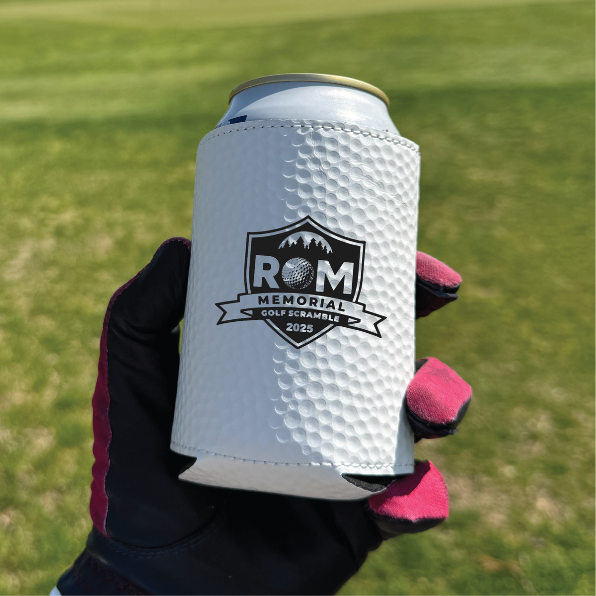 White golf ball-textured can cooler with black shield logo reading "ROM MEMORIAL GOLF SCRAMBLE 2025" and magnetic backing, held in gloved hands on golf course grass