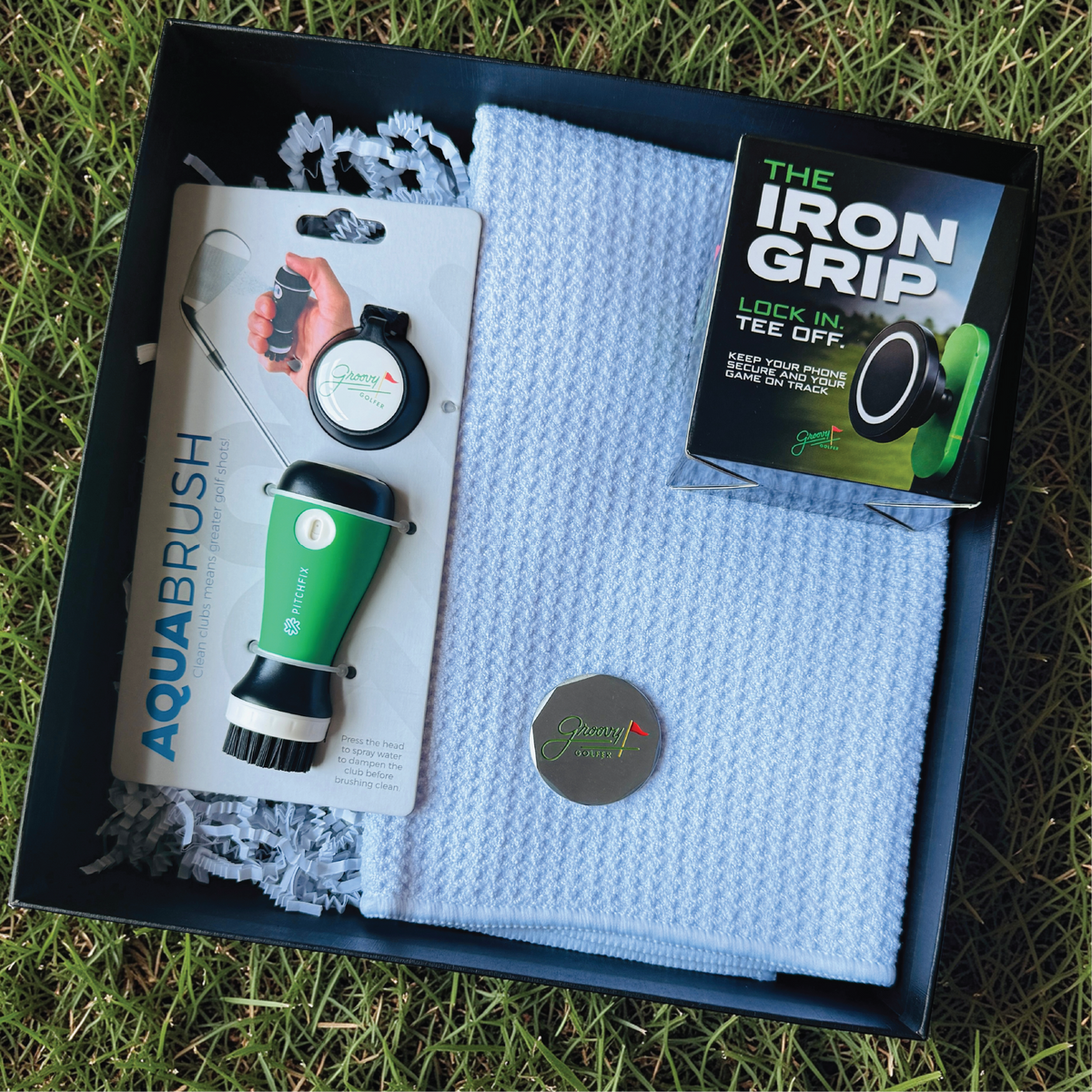 Flat lay of the open golfer's gift box showing the AquaBrush club cleaner, blue waffle towel, magnetic phone mount and golf ball marker.