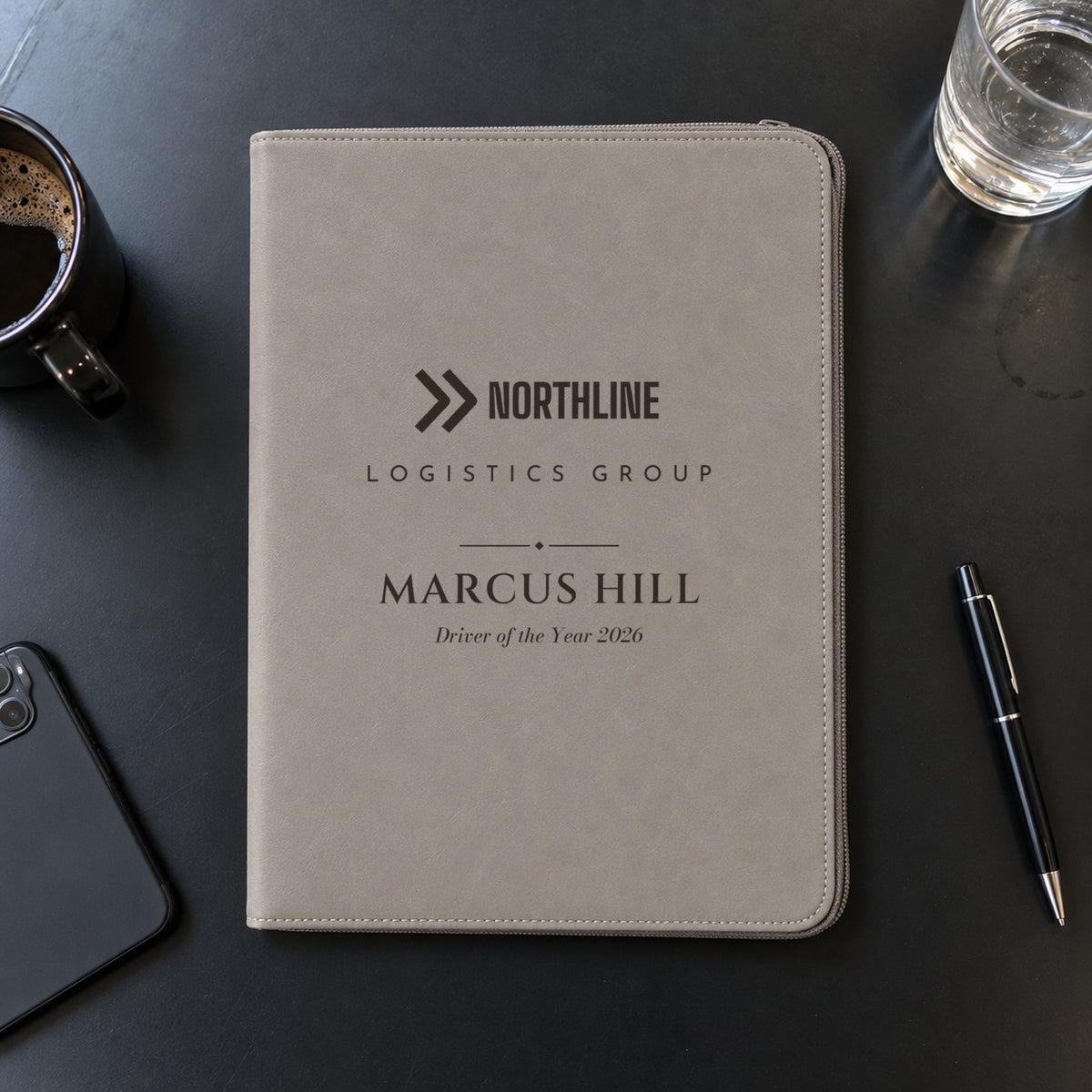 Gray engraved logo padfolio with a sample Northline Logistics logo, Marcus Hill and Driver of the Year 2026 on a black conference table