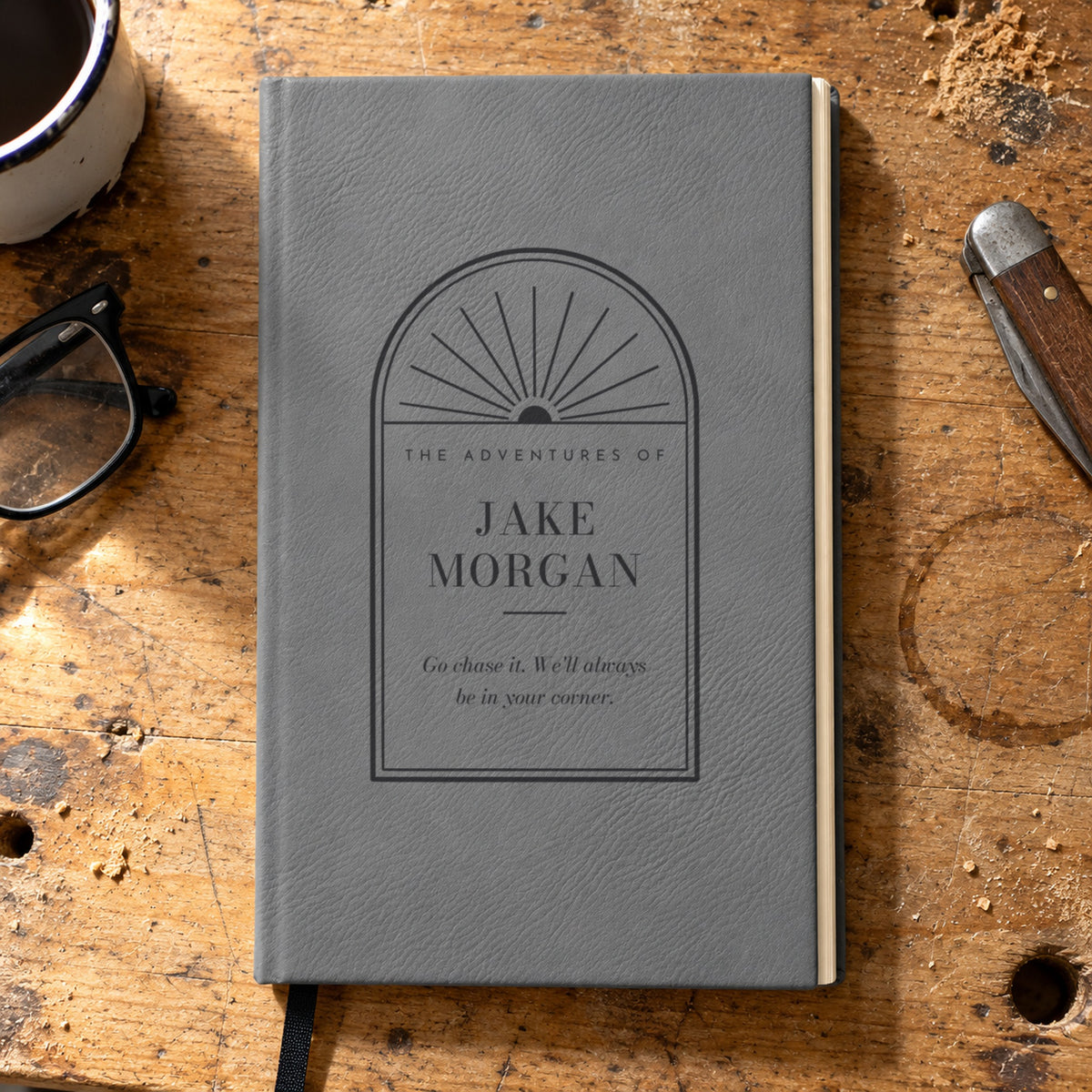 Gray personalized journal engraved The Adventures of Jake Morgan with a message from his parents, a graduation gift for him on a desk