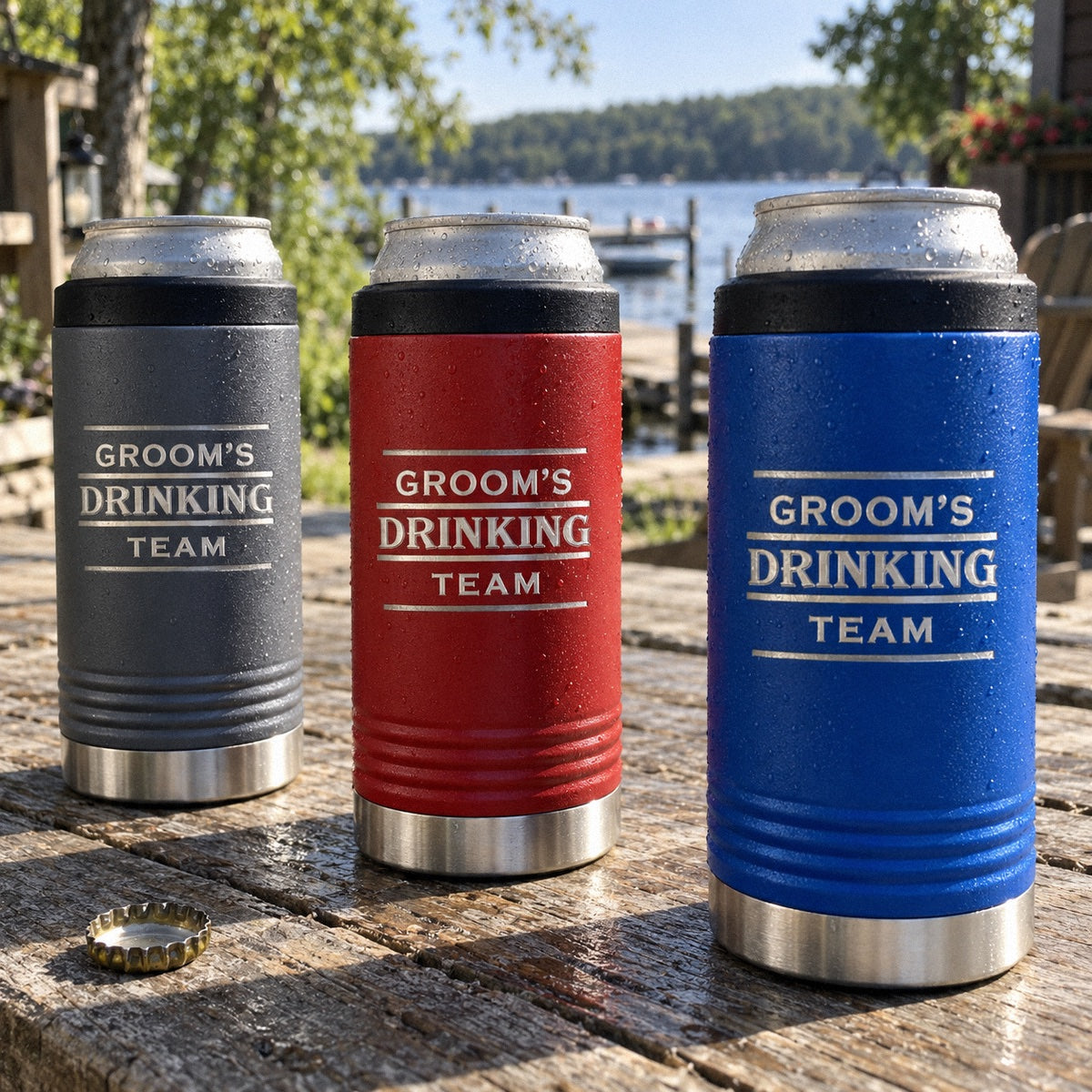 Three slim can coolers engraved GROOM'S DRINKING TEAM in gray, red and blue on a lake house picnic table, a bachelor party gift