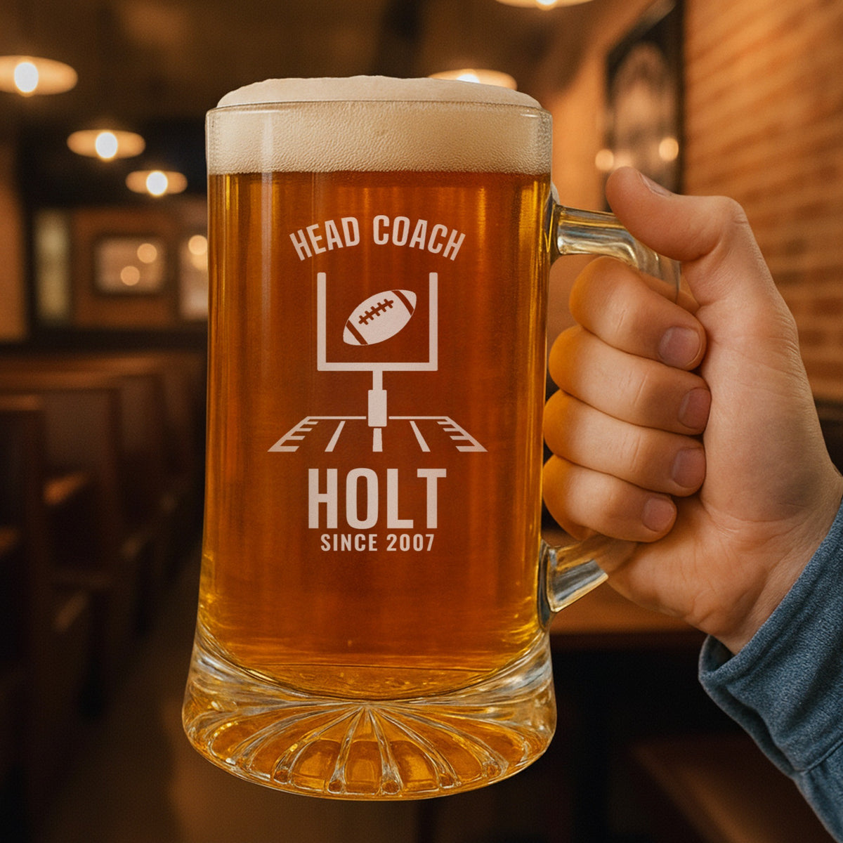 Hand holding the 25 oz personalized head coach football beer stein in a pub, engraved with football goalposts with a ball sailing through: Head Coach, Holt, Since 2007