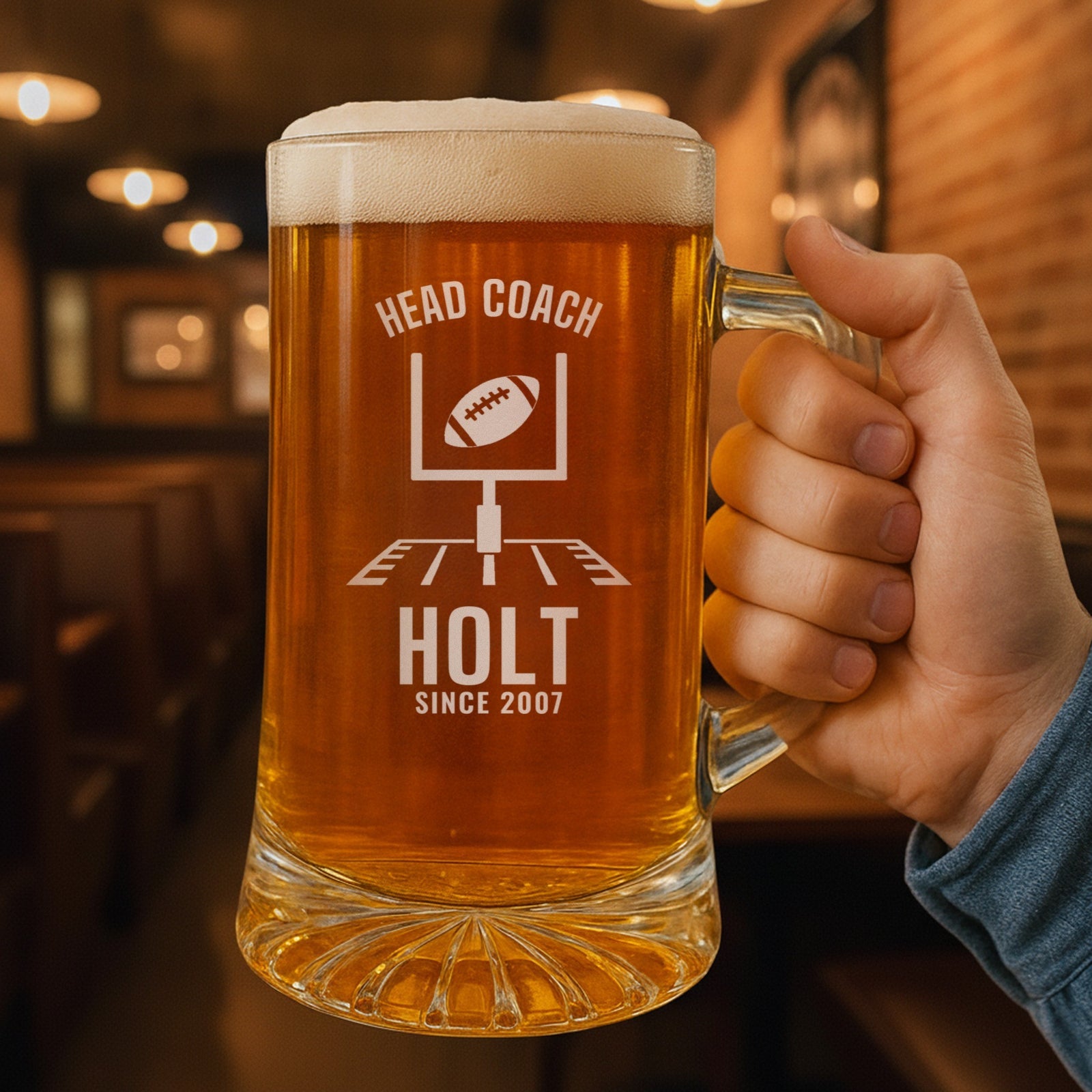 Hand holding the 25 oz personalized head coach football beer stein in a pub, engraved with football goalposts with a ball sailing through: Head Coach, Holt, Since 2007