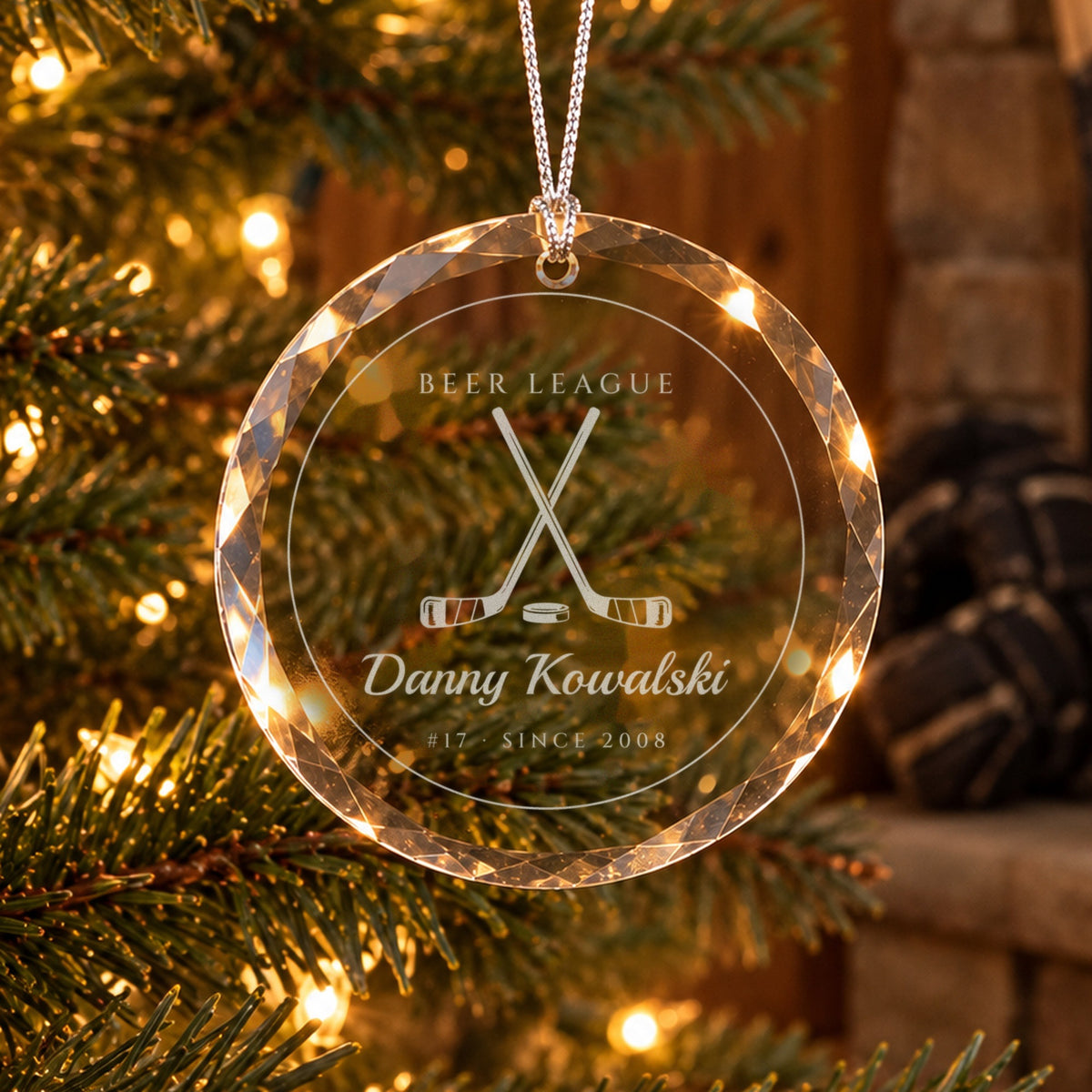 Personalized hockey Christmas ornament in faceted glass on a tree in a wood-paneled den, engraved with crossed sticks and a puck, BEER LEAGUE, Danny Kowalski, #17 · SINCE 2008