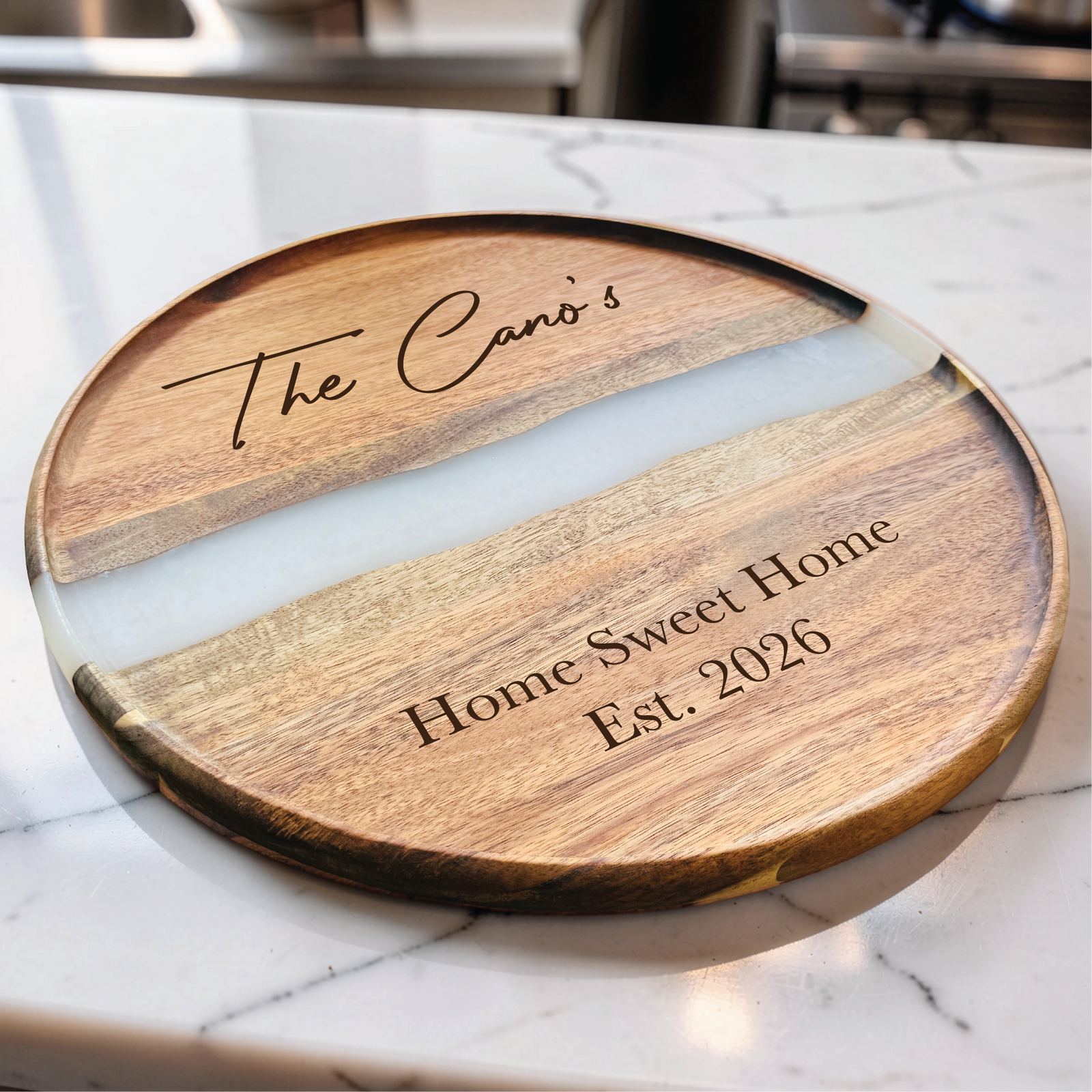 Angled view of the round acacia board with a white resin river, an engraved family name and established date, resting on a marble counter