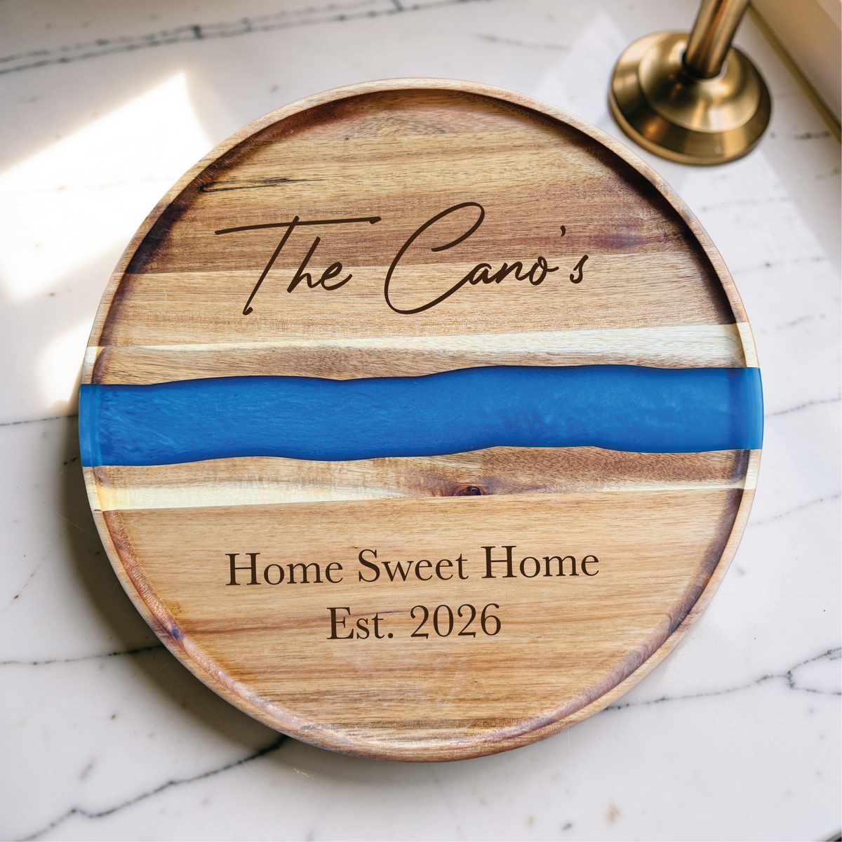 Engraved cutting board with a blue resin river, a script family name above it and Home Sweet Home Est. 2026 below, on a marble countertop