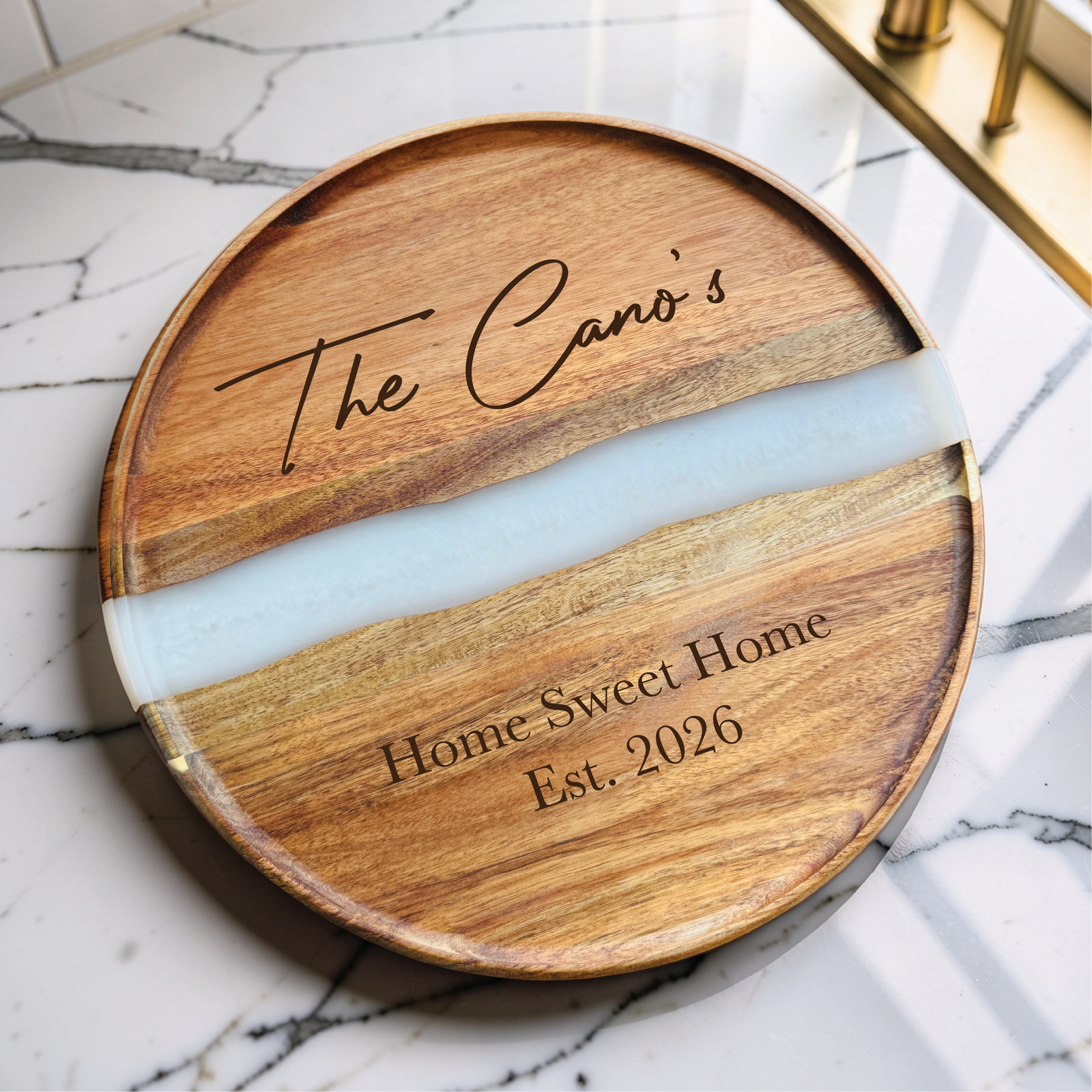 Round acacia board with a clear resin river, engraved with a script family name and Home Sweet Home Est. 2026, on a marble counter