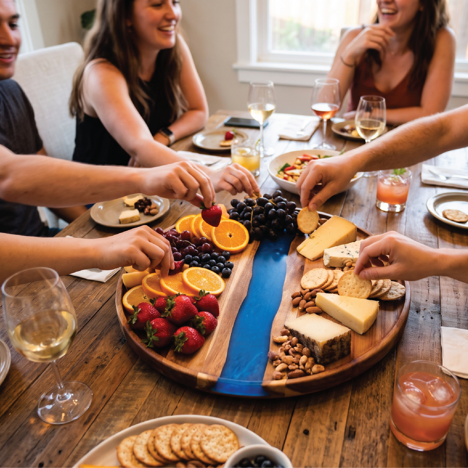 Friends reaching for fruit, cheese and crackers served on a round acacia board with a blue resin river at a dinner table with wine