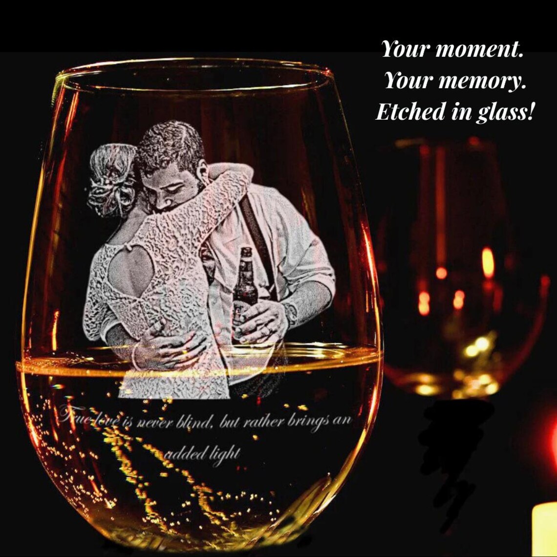 Stemless wine glass filled with amber liquid, laser-engraved with sample photo of couple embracing and decorative text, shown against candlelit background