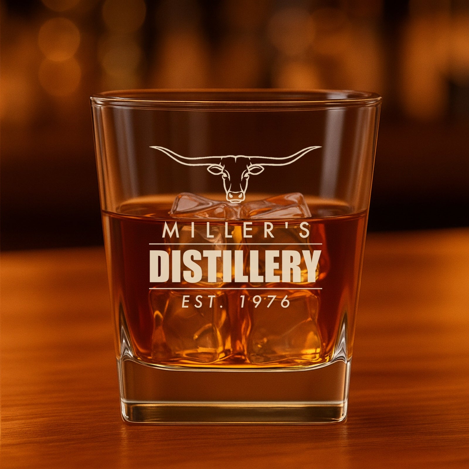 Personalized whiskey glass engraved with a longhorn steer over Miller's, Distillery and Est. 1976, filled with bourbon on a bar top