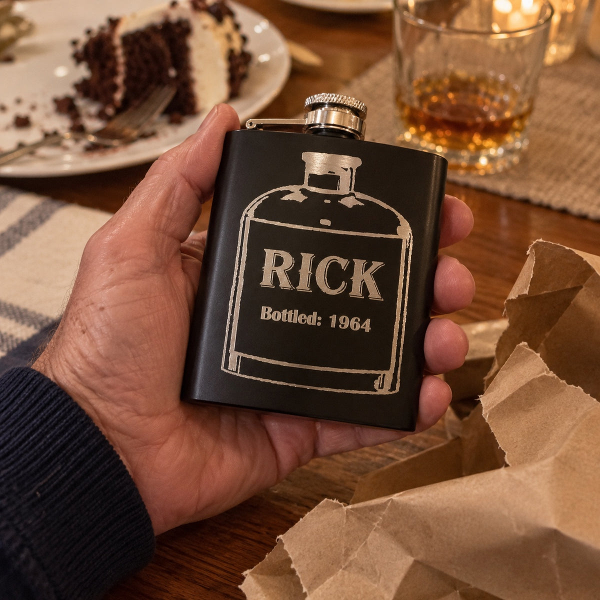 A man holds his engraved flask reading RICK and Bottled: 1964 beside torn kraft paper and birthday cake, a thoughtful birthday gift for men