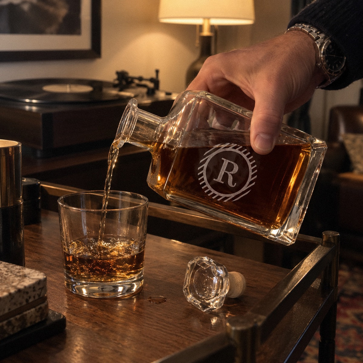 A man pours whiskey from a monogrammed decanter etched R into a rocks glass on his bar cart, a personalized whiskey gift for him