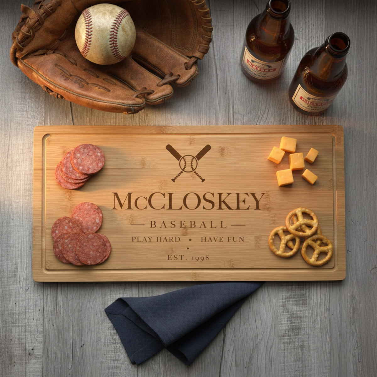 Engraved baseball bamboo board on a weathered wood table with sliced summer sausage at one end, cheddar and pretzels at the other, beside a glove and two beers.