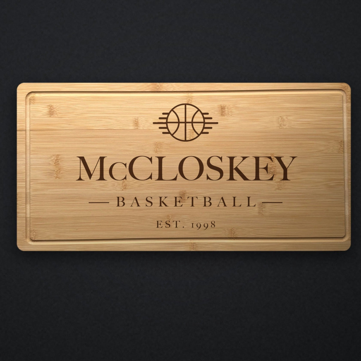 Personalized basketball cutting board in solid bamboo on dark slate, engraved with a basketball emblem above the family name and the established year.