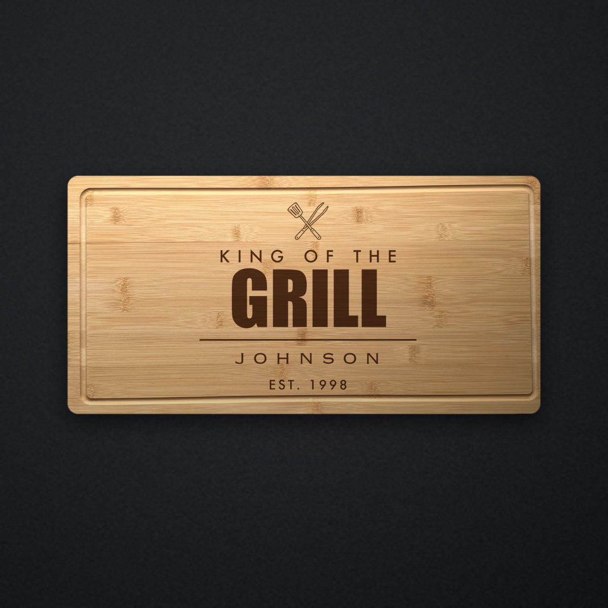 Personalized BBQ cutting board in solid bamboo on dark slate, engraved King of the Grill above the family name and the established year.