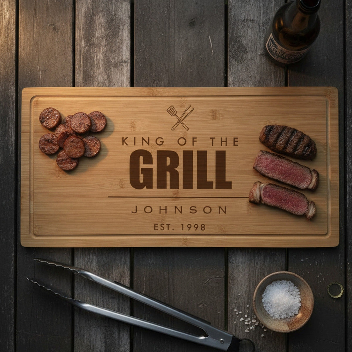 Engraved King of the Grill bamboo board on a dark outdoor table with sliced grilled sausage at one end, sliced ribeye at the other and tongs beside it.