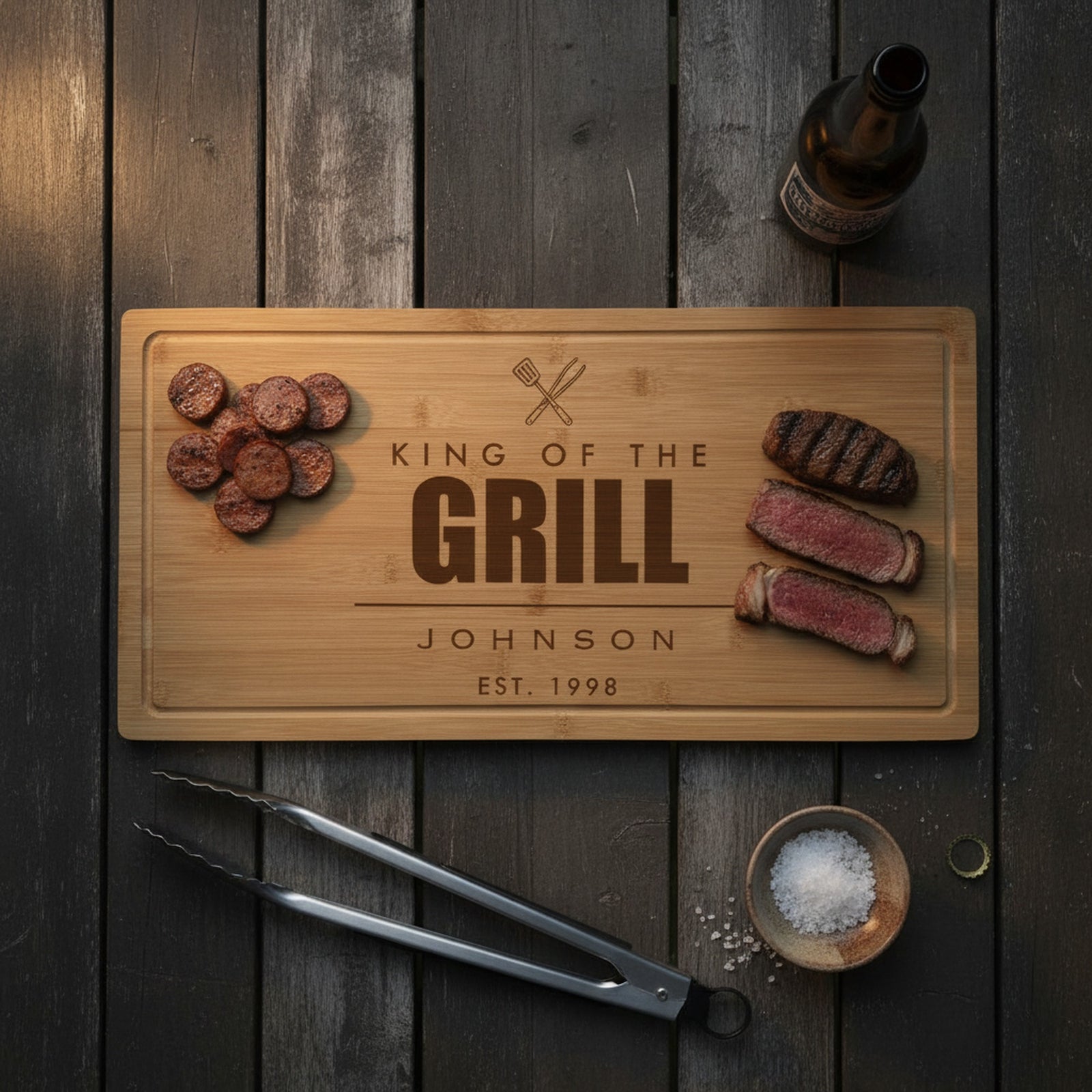 Engraved King of the Grill bamboo board on a dark outdoor table with sliced grilled sausage at one end, sliced ribeye at the other and tongs beside it.