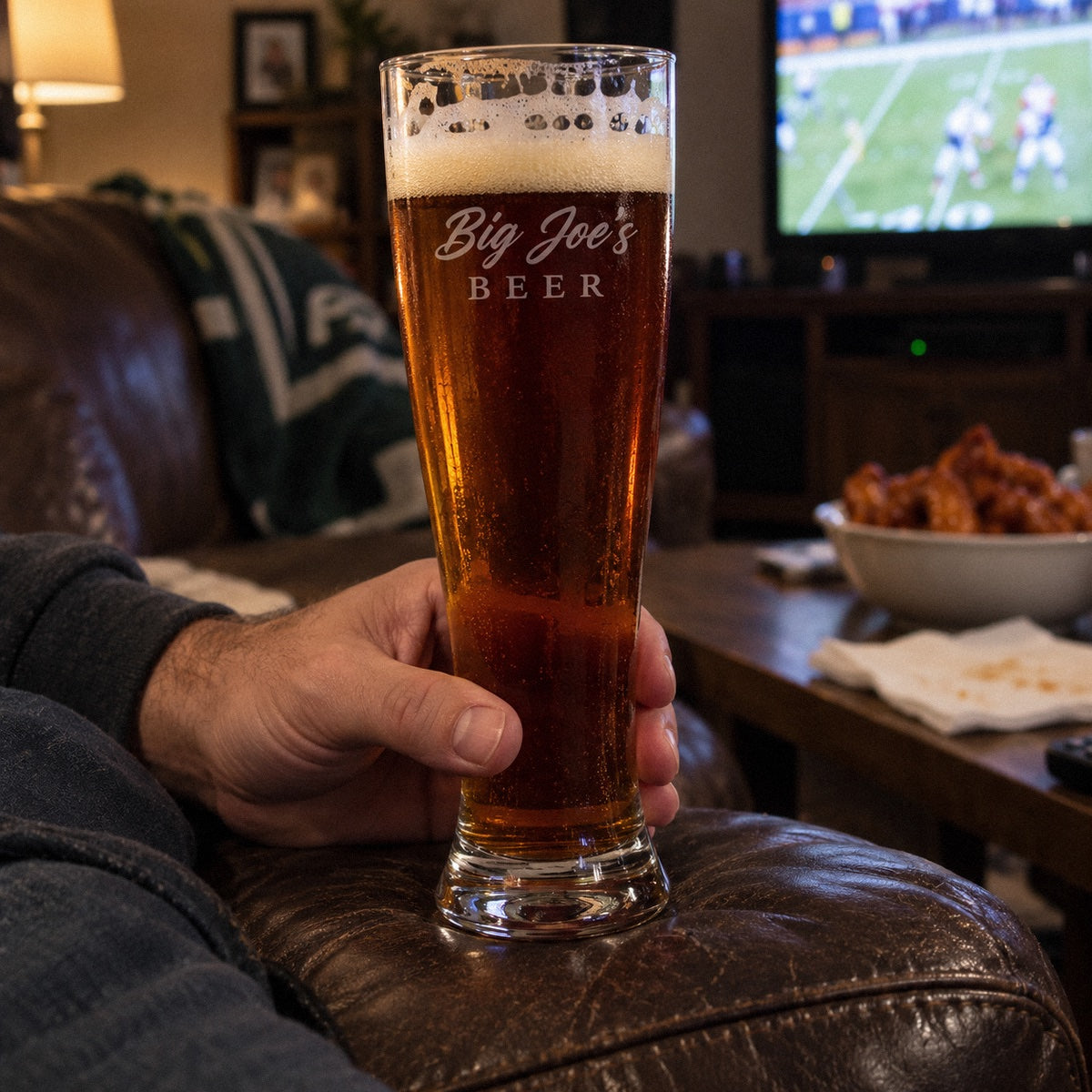 A man holds a tall personalized beer glass etched Big Joe's BEER on the couch arm on game day, an engraved beer glass gift for him