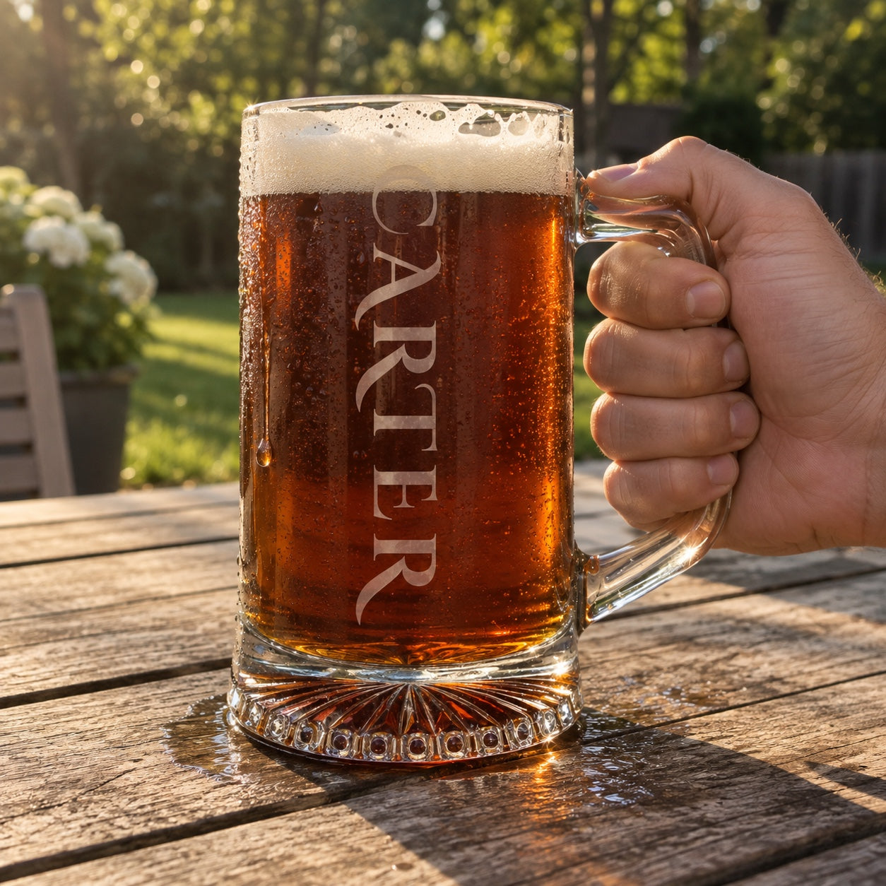 Personalized beer stein engraved CARTER down the side in frosted capitals, a heavy glass stein of amber ale held on a backyard patio table