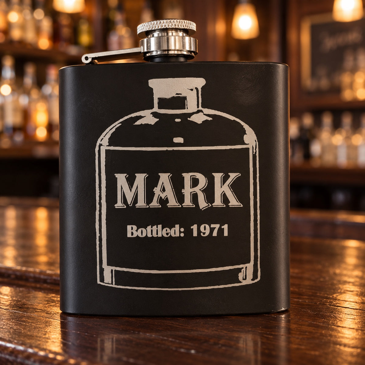 Matte black birthday flask on a dark wood bar, engraved with an old bottle label reading MARK and Bottled: 1971, a personalized birthday gift for him