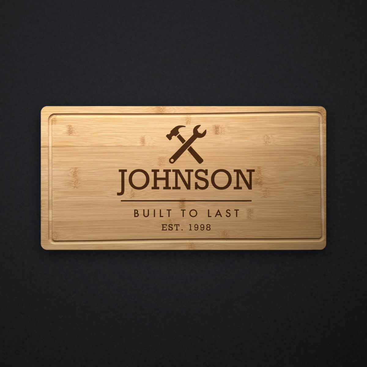 Personalized blue collar cutting board in solid bamboo on dark slate, engraved with a crossed hammer and wrench above the family name and Built to Last.