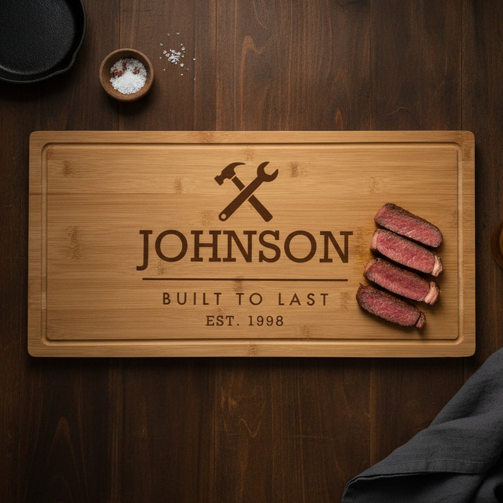 Engraved Built to Last bamboo steak board on a dark walnut table with sliced ribeye at one end, a cast iron skillet, sea salt and a glass of beer.
