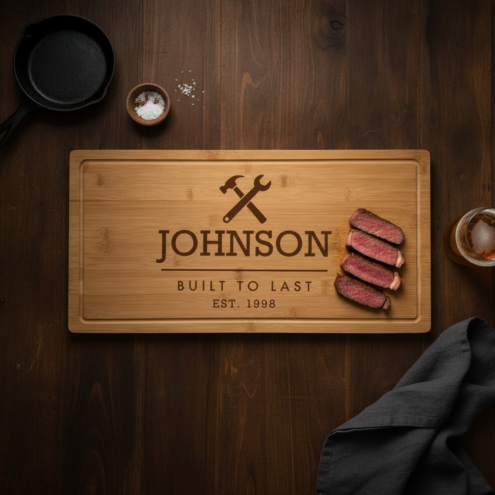 Engraved Built to Last bamboo steak board on a dark walnut table with sliced ribeye at one end, a cast iron skillet, sea salt and a glass of beer.