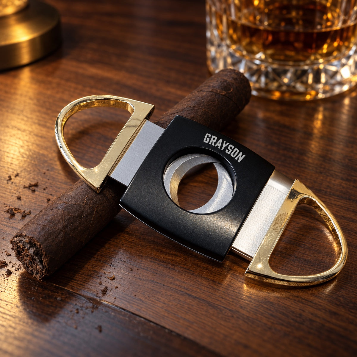 Personalized cigar cutter in black and gold engraved GRAYSON, a double blade guillotine cutter lying across a cigar on a walnut table beside a whiskey glass.