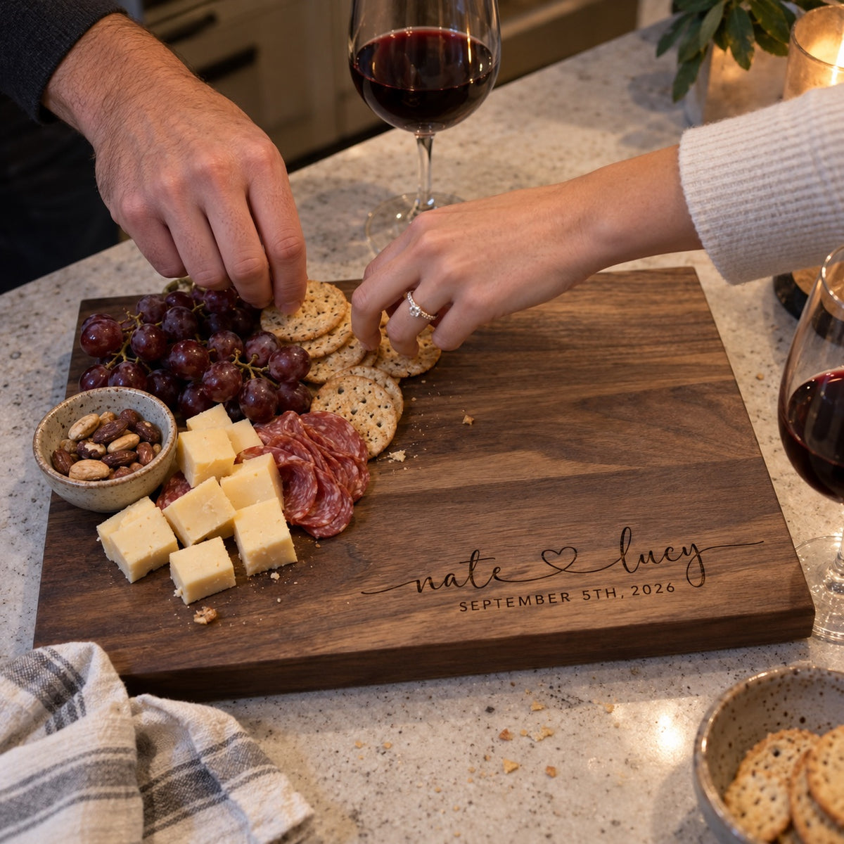 A couple shares crackers, cheese and salami on a walnut cutting board engraved nate and lucy, a personalized anniversary gift for the kitchen