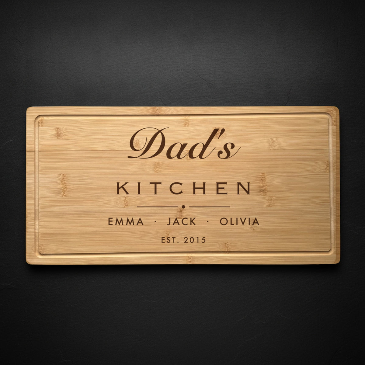 Personalized dad cutting board in solid bamboo on dark slate, engraved Dad's Kitchen in flowing script above the names of his three kids and the year.