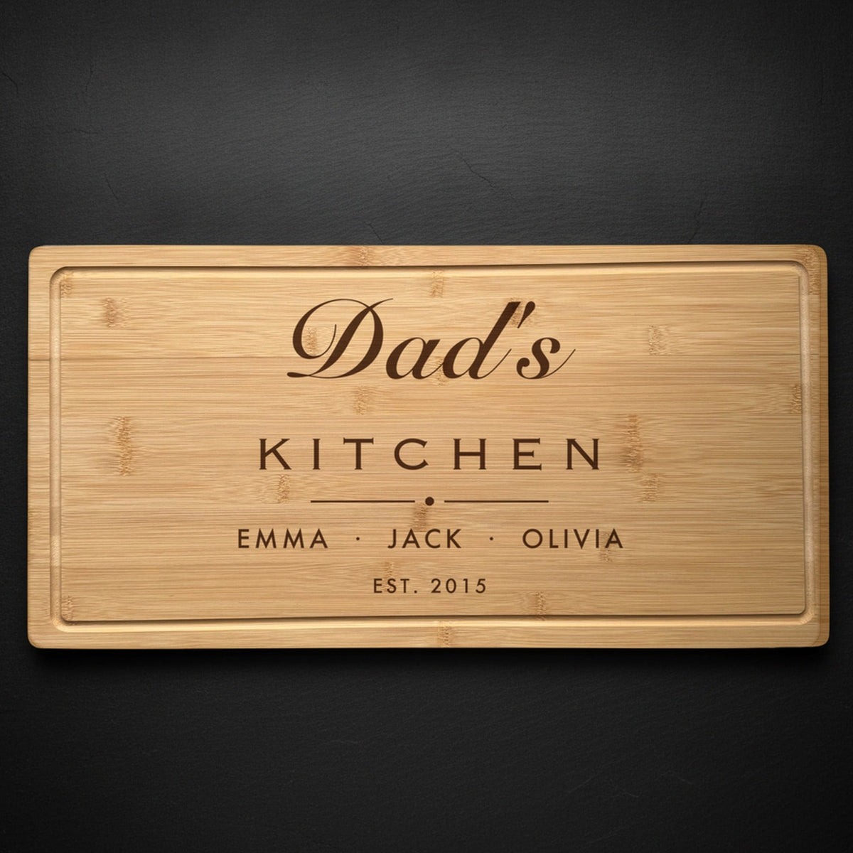 Personalized dad cutting board in solid bamboo on dark slate, engraved Dad's Kitchen in flowing script above the names of his three kids and the year.