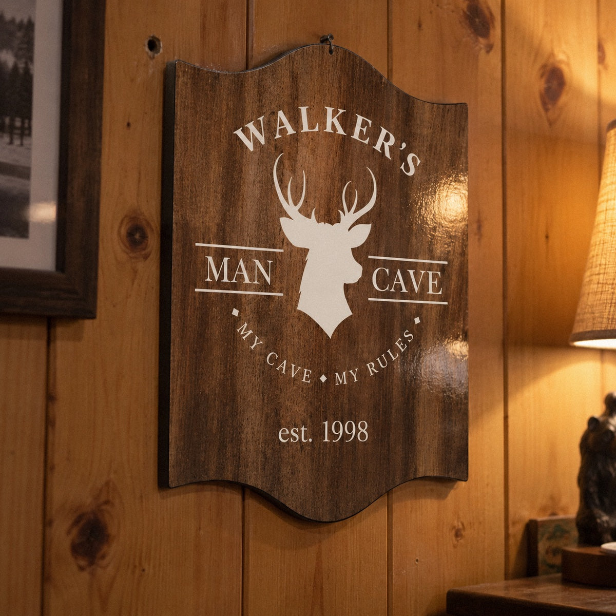 Personalized deer head wall sign printed WALKER'S, MAN CAVE and est. 1998 on a high-gloss wood-look plaque hanging on a knotty pine basement wall