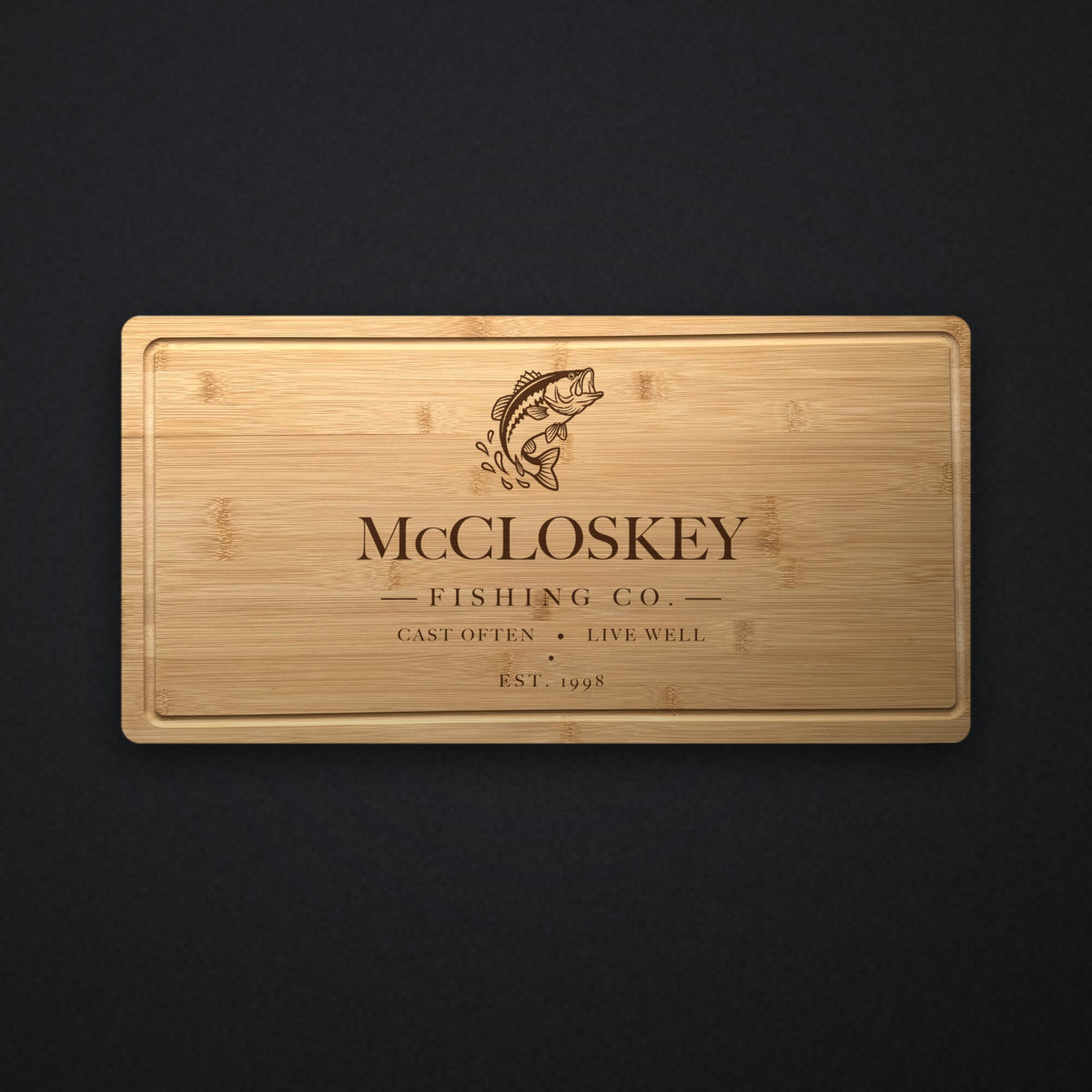 Personalized fishing cutting board in solid bamboo on dark slate, engraved with a leaping bass above the family name, Fishing Co. and the year.