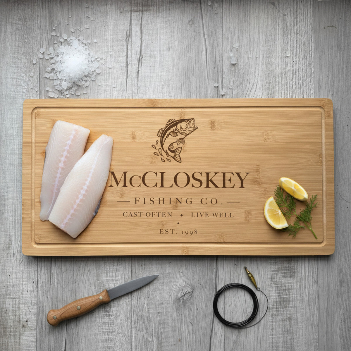 Engraved fishing bamboo board on weathered dock planking with two fresh fish fillets at one end, lemon wedges and dill at the other, beside a fillet knife.