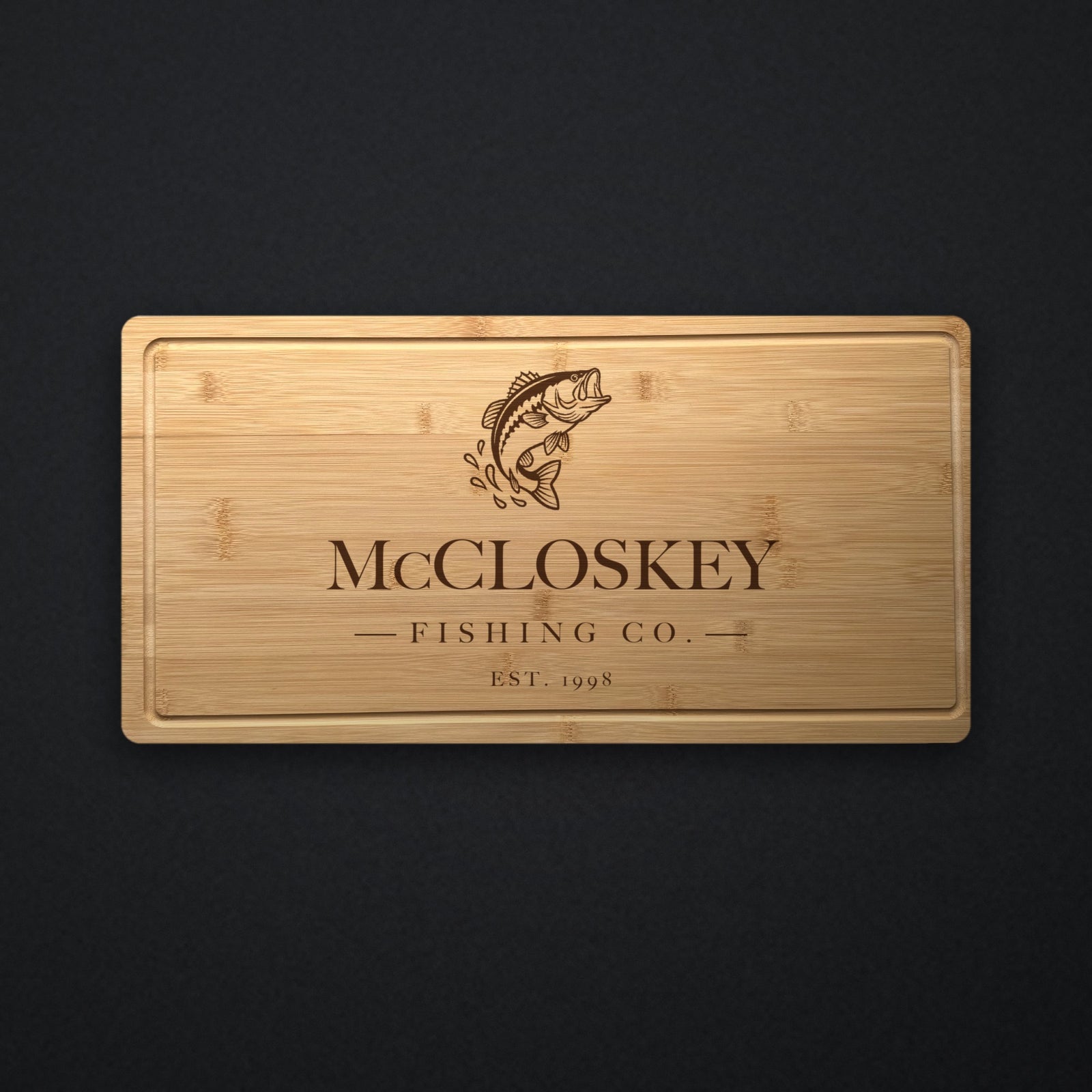 Personalized fishing cutting board in solid bamboo on dark slate, engraved with a leaping bass above the family name, Fishing Co. and the year.