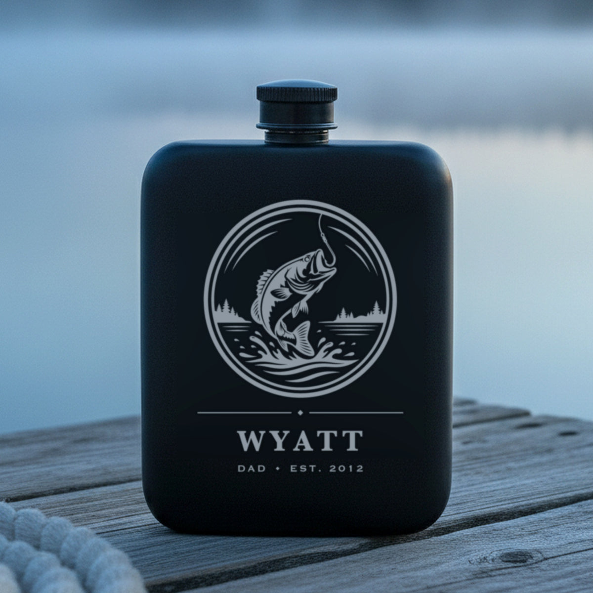 Personalized bass engraved fishing flask standing on the weathered boards of a lake dock at first light with mist over flat water.