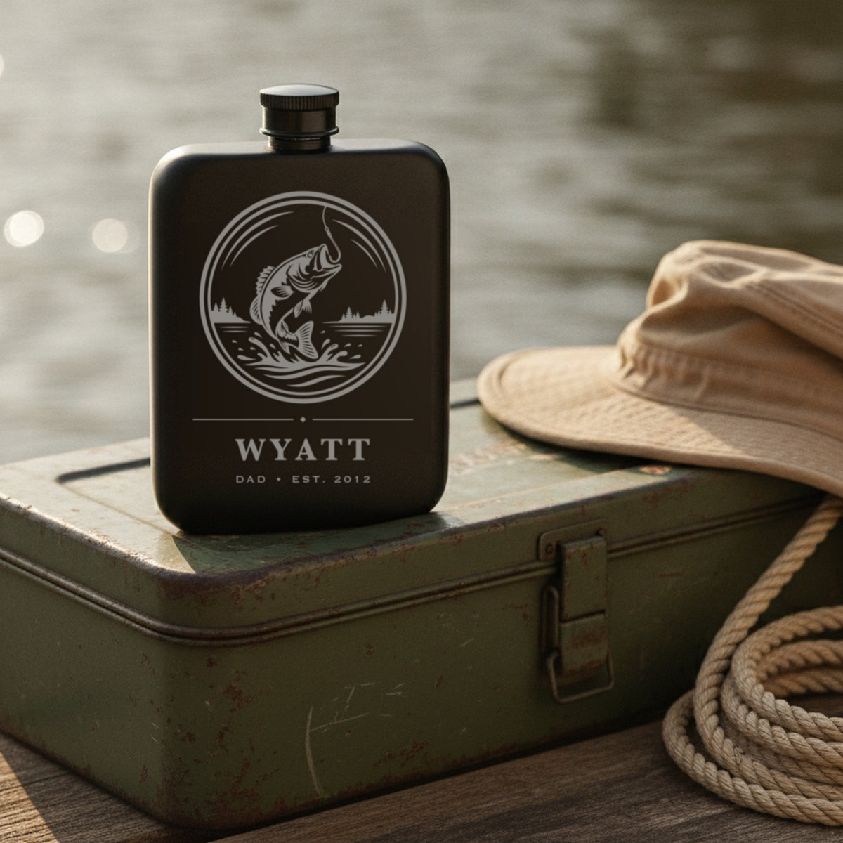 Engraved fishing flask on the lid of a worn green tackle box beside a canvas hat and a coil of rope on a sunny dock.