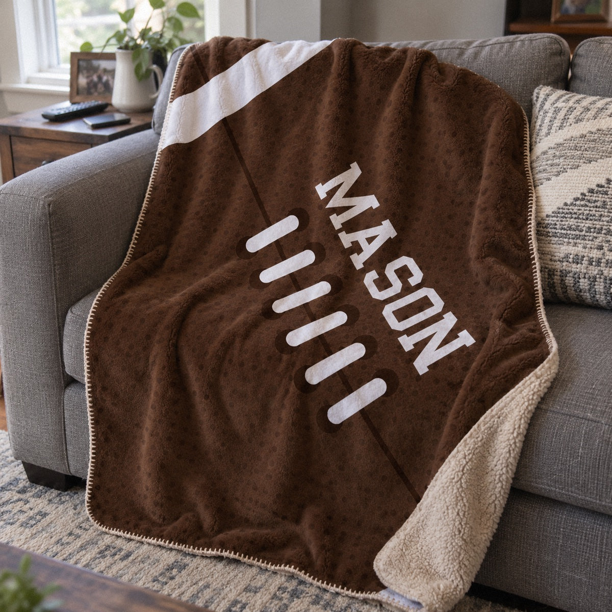 Personalized football sherpa blanket printed like a football with the name MASON along the laces, draped over a gray couch in a family room