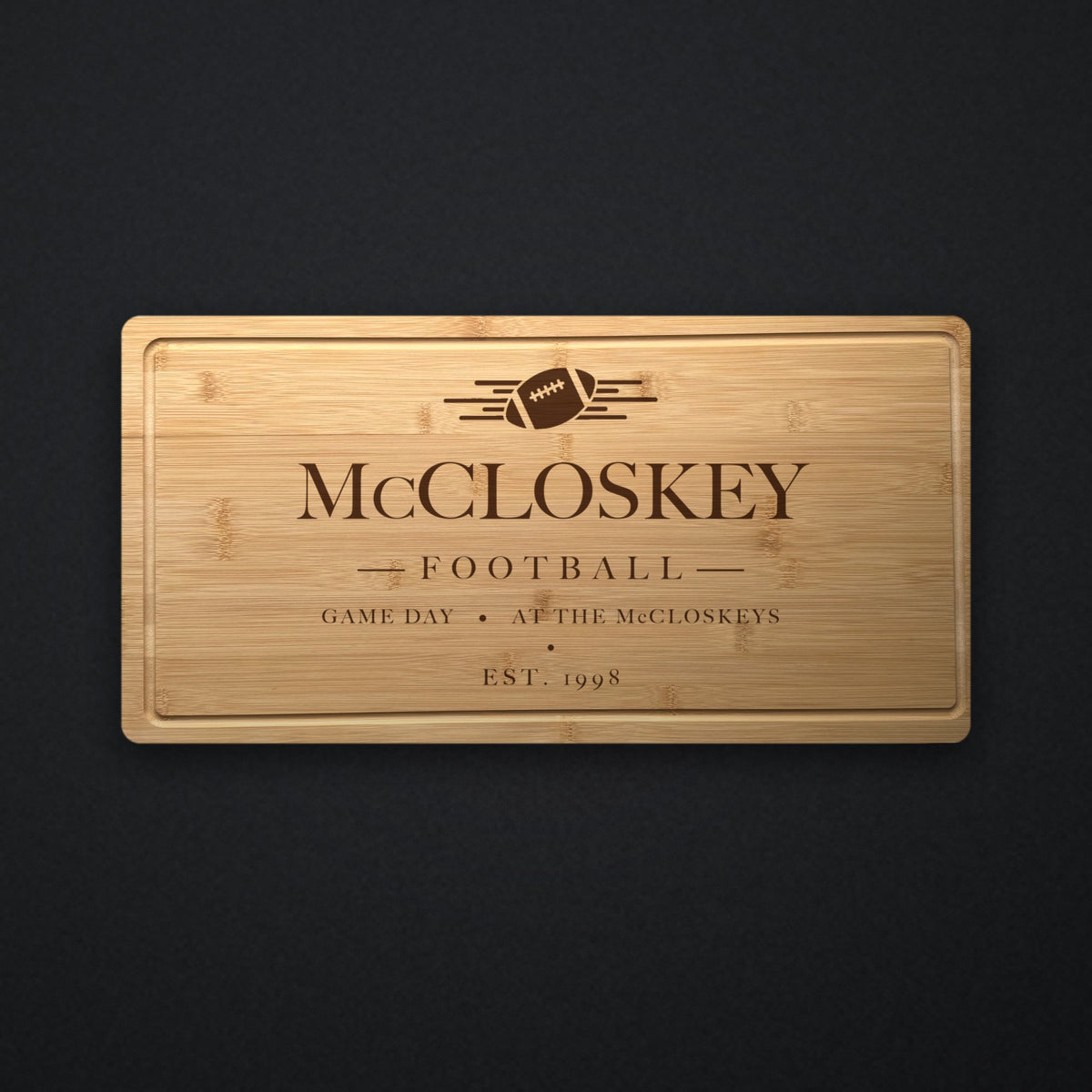 Personalized football tailgate cutting board in solid bamboo on dark slate, engraved with a football and speed lines above the family name and the year.