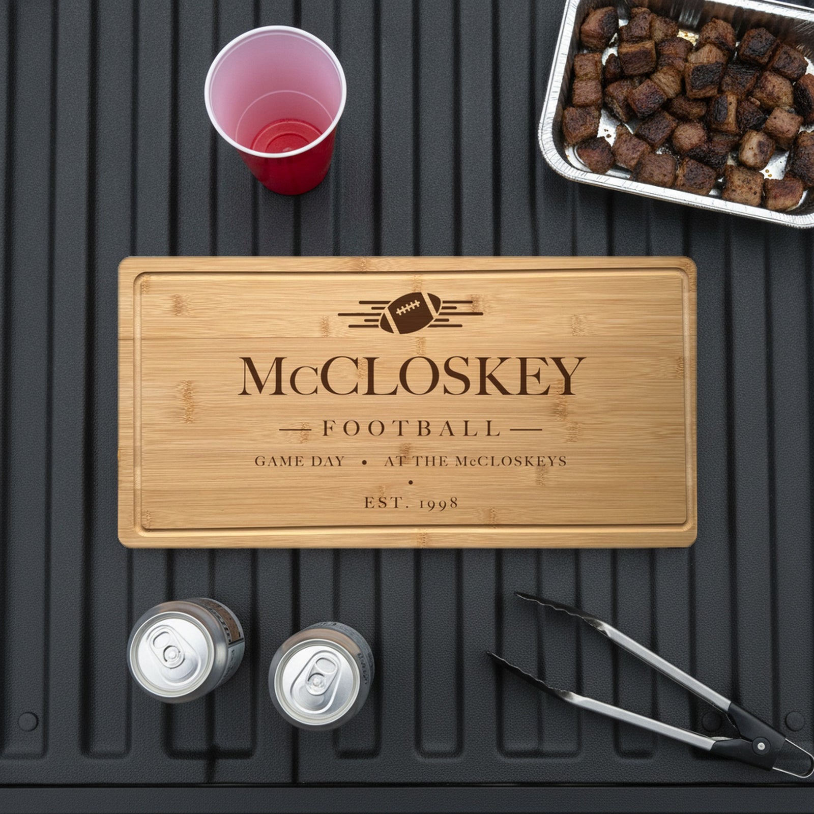 Engraved football bamboo board on the open tailgate of a pickup truck beside a tray of grilled burnt ends, two cans, a red cup and grill tongs.