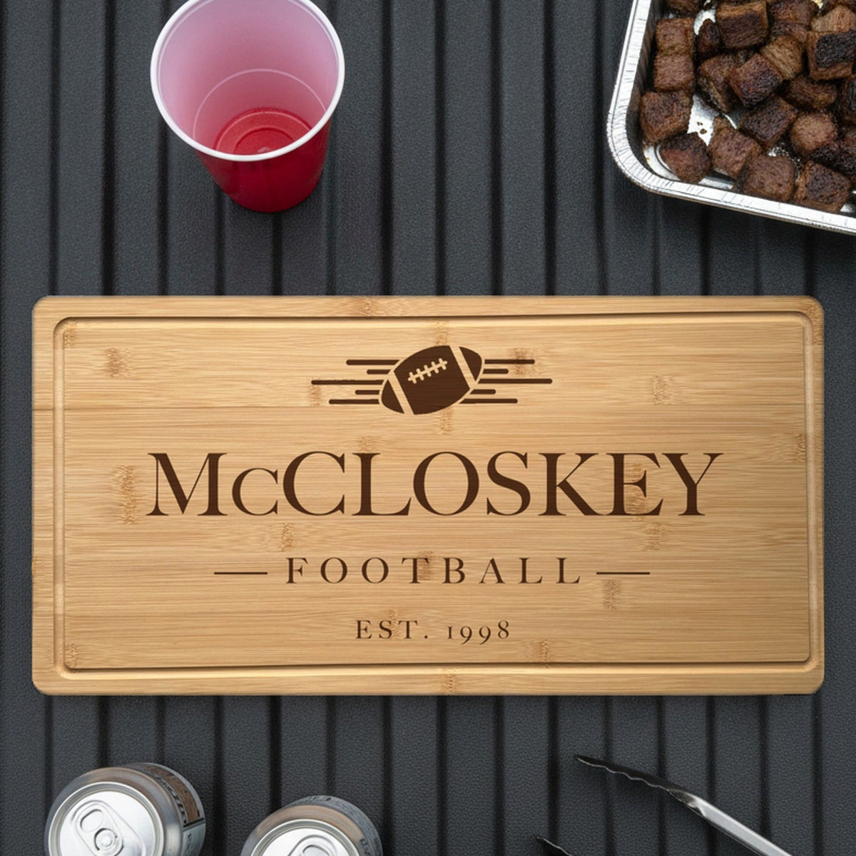 Engraved football bamboo board on the open tailgate of a pickup truck beside a tray of grilled burnt ends, two cans, a red cup and grill tongs.