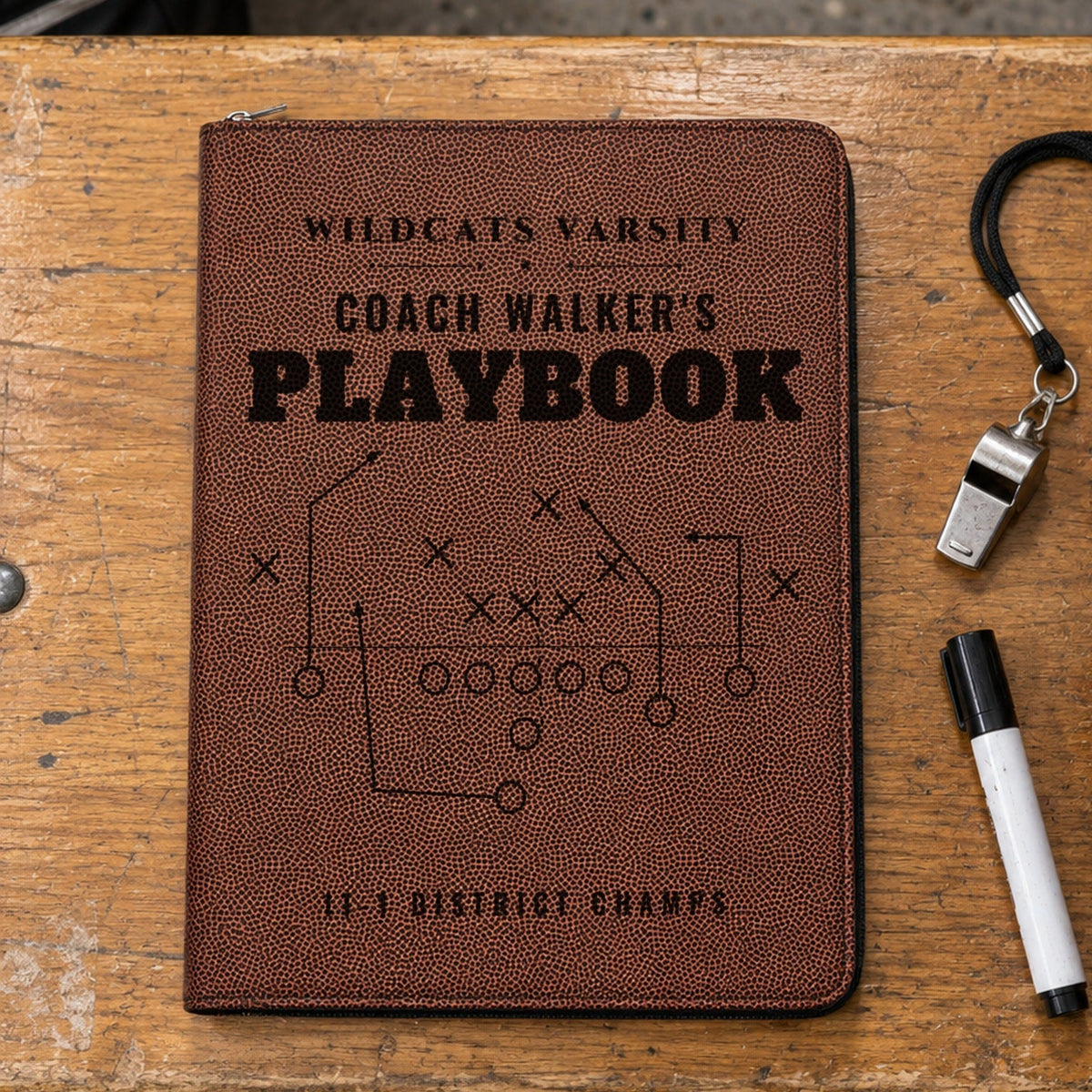 Personalized coach portfolio in football grain leather engraved Wildcats Varsity, Coach Walker's Playbook, 11-1 District Champs, on a locker room bench beside a whistle